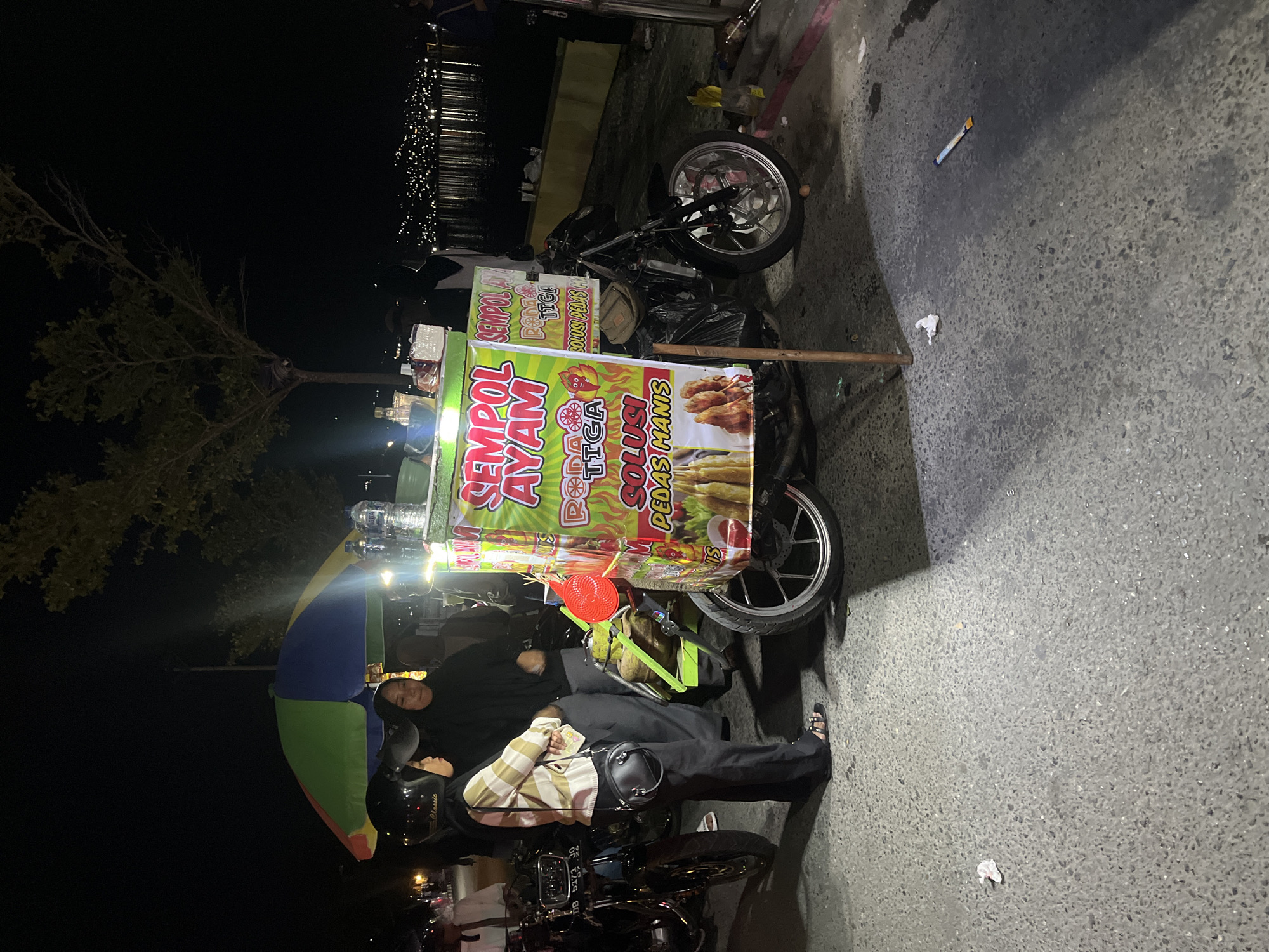 At night, a food cart selling "Sempul Ayam" is parked on a paved area next to a body of water, with lights from a distant city visible across the water, likely in Luwuk, Indonesia. Two women, one wearing a hijab and a striped long-sleeved shirt, and another in a black hijab, stand near the cart. The cart itself is adorned with colorful banners advertising the food, featuring images of chicken skewers. A motorcycle is parked to the right of the cart. The ground is covered in asphalt, with some debris scattered around, including a small piece of paper and what appears to be a cigarette butt. Trees are visible in the background, and the overall scene suggests a street-side food stall operating in the evening.