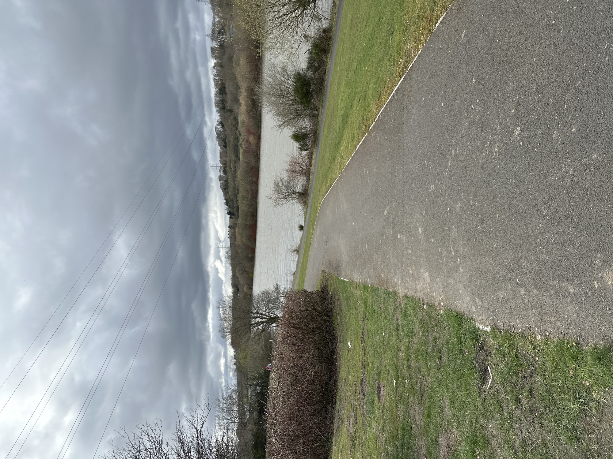 The image depicts a path alongside a body of water, likely a lake or reservoir, near Dunfermline, United Kingdom. The path is made of asphalt and borders a grassy area on one side and the water's edge on the other. The water appears choppy, possibly indicating wind. Beyond the water are trees and vegetation. Overhead, power lines are visible against a cloudy sky.