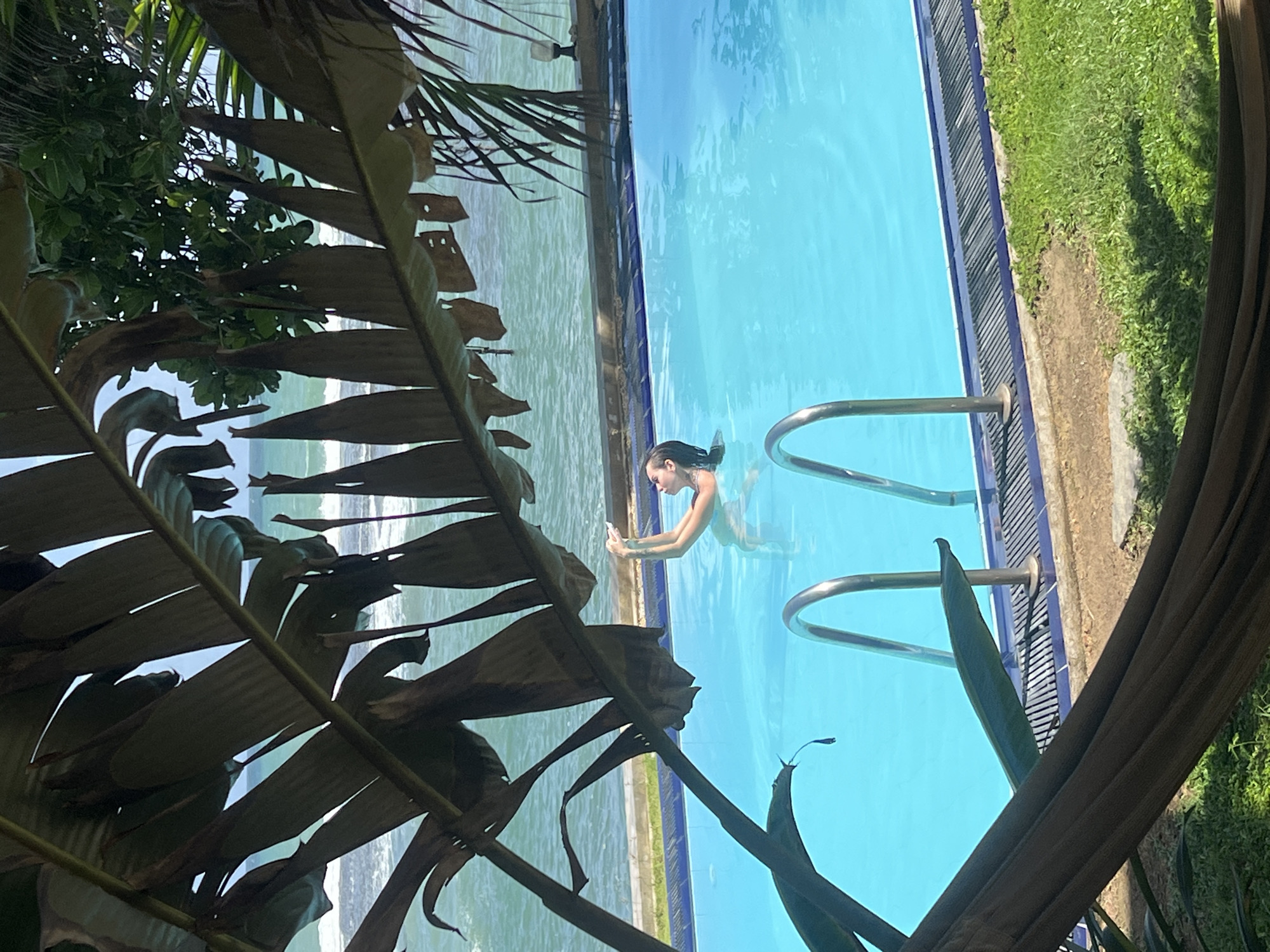 In the city of Goviyapana, Sri Lanka, a person is submerged in a bright blue swimming pool, their arms raised above their head. The pool is bordered by a tiled edge and a metallic railing. The background features the ocean with gentle waves and a distant shoreline. Large, dark green leaves from a tropical plant overhang the scene from the foreground, casting shadows and framing the pool and ocean. The ground beside the pool is covered in green grass.