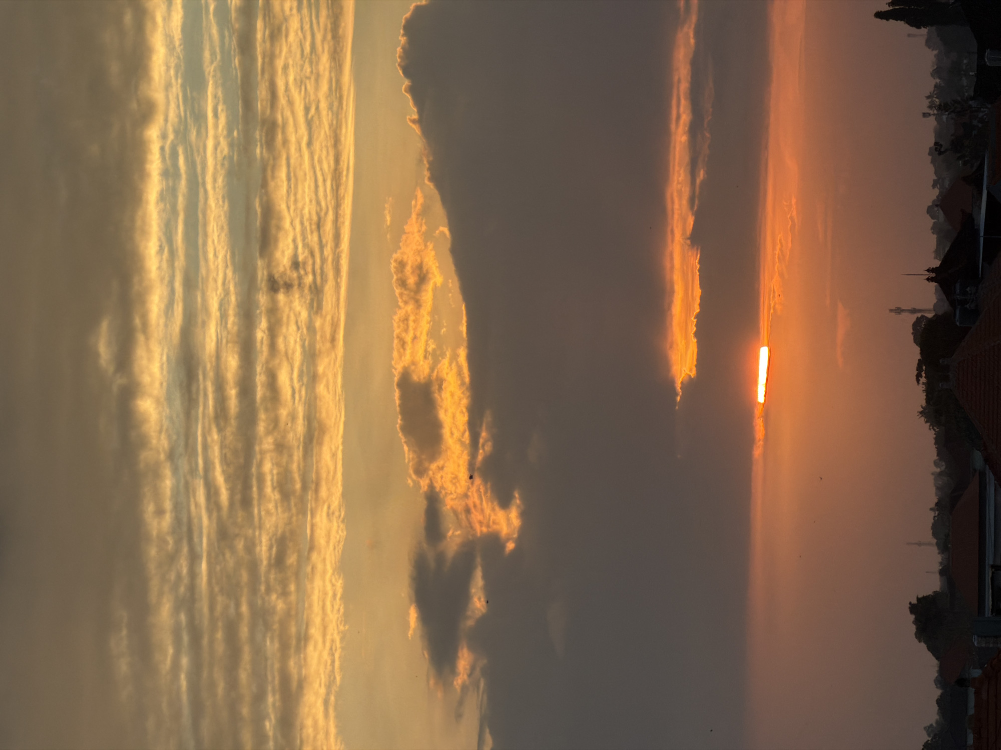 The image captures a sunset in North Kuta, Indonesia. The sky is dominated by orange and yellow hues. A large, dark cloud formation is visible in the middle, partially obscuring the setting sun. The sun appears as a bright, elongated line through a gap in the clouds. To the right, rooftops and vegetation indicate a residential area. Several small dark spots, likely birds, are scattered across the sky.