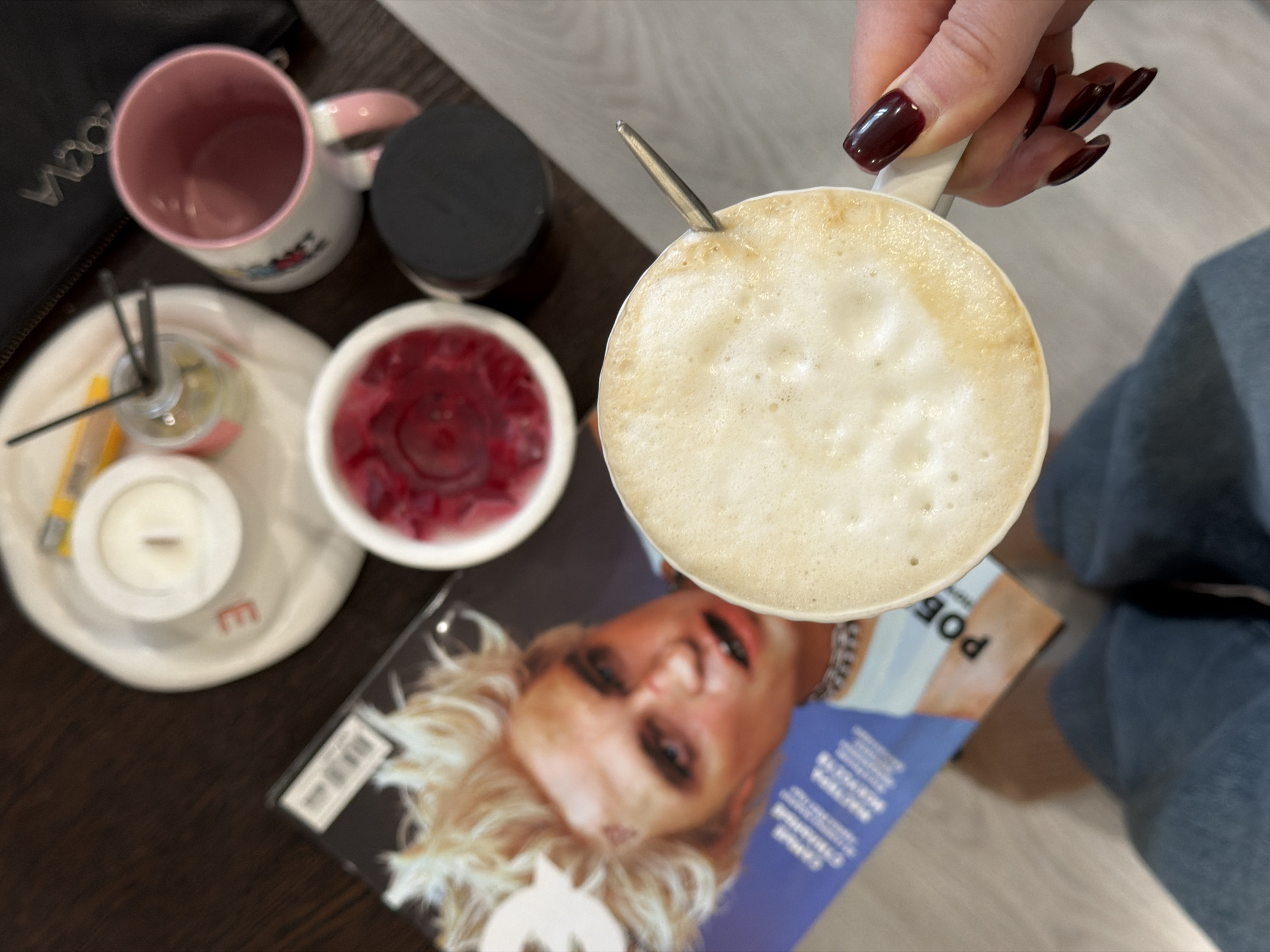 This image, captured from a high-angle, provides a close-up view of a person holding a cup of a frothy beverage, likely coffee or a latte, within an indoor setting in Засечное, Russia.
The central subject is a person's hand, adorned with neatly manicured nails painted a dark red or burgundy color, gripping a white mug. A small metal spoon is inserted into the creamy, textured foam of the drink. Partially visible in the lower right is the person's leg, clad in blue jeans.
Arranged on a dark tabletop with lighter flooring visible, various objects contribute to a cozy and relaxed atmosphere. To the left of the main cup, a white mug with a pink rim, possibly featuring cartoon-like decorations, sits near a black bag or purse where the partial text "VOOM" is visible. A small white tray holds a reed diffuser with dark reeds, a small white candle in a holder (with its wick showing), and a yellow object. Adjacent to the tray is a white bowl filled with a red, jelly-like substance or berries, possibly a dessert. A black lidded container is also present.
Underneath the main cup and other items, a magazine lies open, with its cover featuring a close-up of a blonde woman's face, strongly resembling the artist Madonna, printed upside down from the viewer's perspective. Visible partial Cyrillic text on the magazine includes "ПОБ" (potentially part of "Победа") and other characters. A small letter "e" is also visible on the white tray.
The overall scene suggests a moment of leisure, possibly a coffee break or a relaxing personal moment. The combination of a warm drink, decorative elements like the candle and diffuser, and a magazine implies comfort and tranquility.
