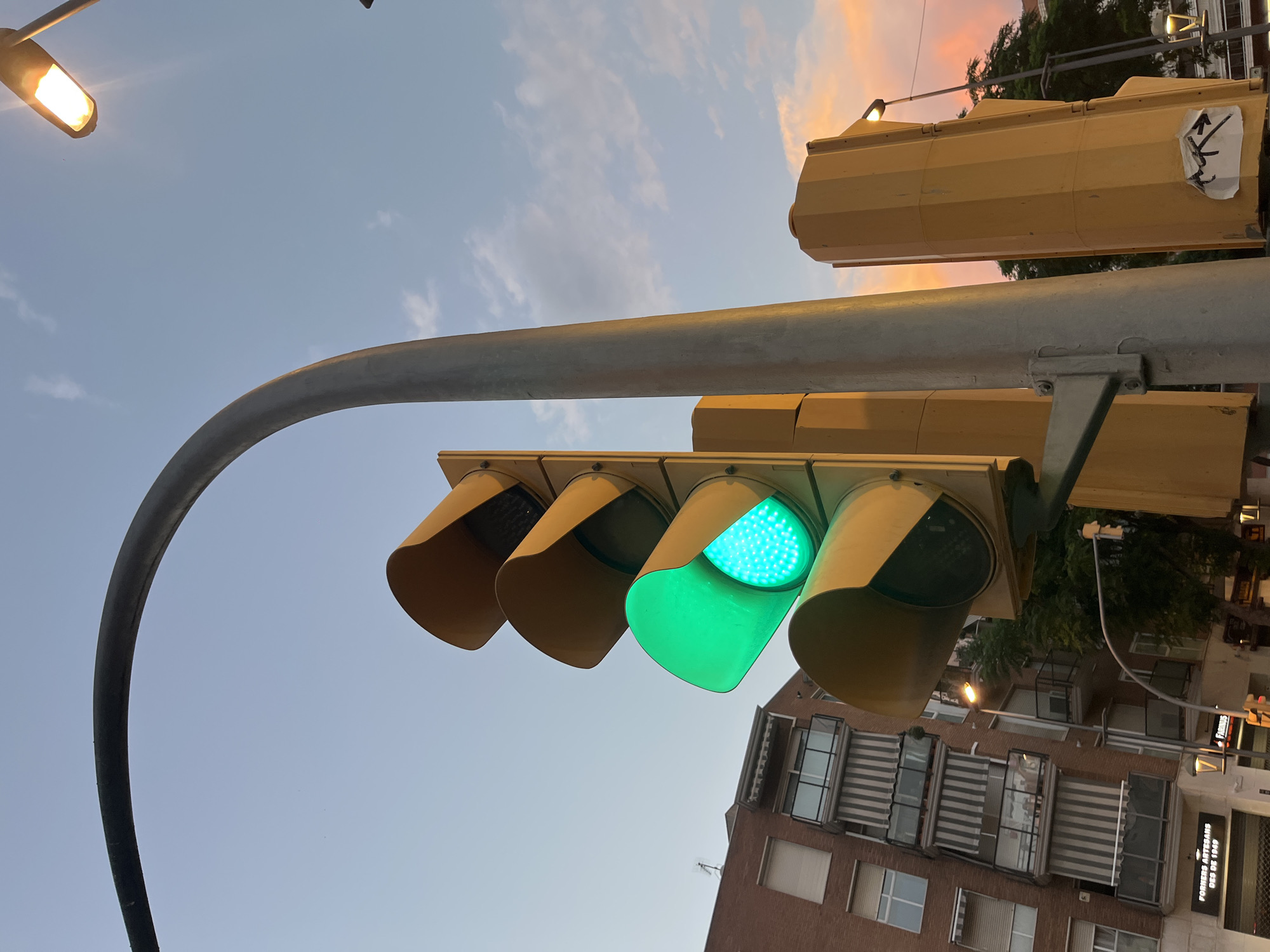 The image captures a close-up, low-angle view of a traffic light in Tarragona, Spain, with the green light illuminated. The traffic light, predominantly yellow, is mounted on a grey pole. Above and to the right, a street lamp with a warm, orange glow is visible against a dusky sky with soft pink and blue clouds. In the background, a multi-story brick building with balconies featuring striped awnings is present, suggesting an urban setting. The lower portion of the frame shows a street sign with text that is partially obscured but appears to read "FORNERS ARTESANS DES DE 1949". The overall scene is set at dusk or early evening.