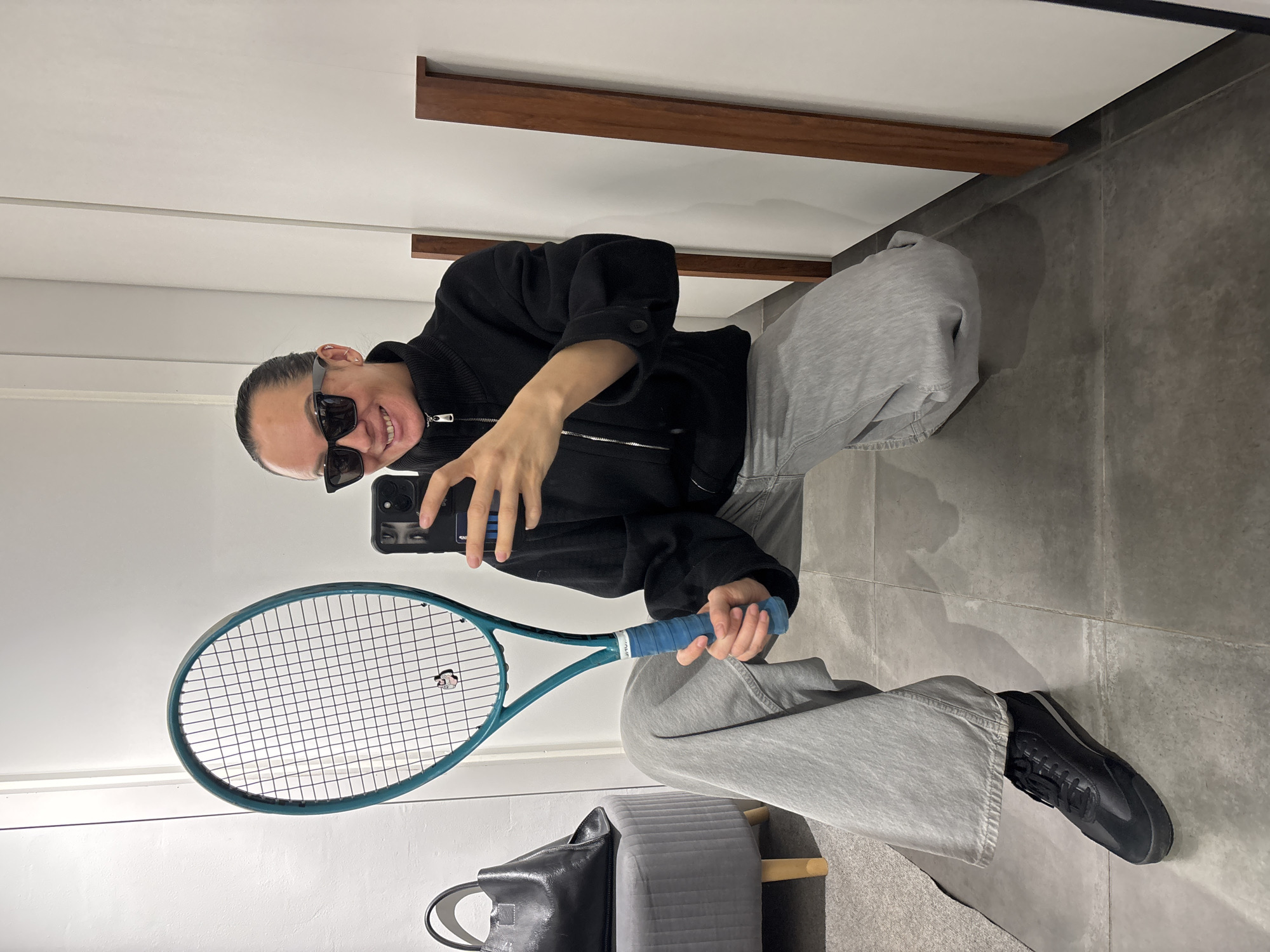 A person is taking a selfie while seated in what appears to be a fitting room or a bathroom, holding a tennis racket. The person is wearing sunglasses, a black zip-up jacket, and gray pants. They are holding a smartphone in their left hand, presumably to take the photo, and the tennis racket in their right hand.
The setting appears to be indoors, likely in a fitting room given the presence of a mirror and a clothes rail. The background consists of a white wall with a wooden clothes rail, and a gray tiled floor. There is a black bag near the bottom of the image and a gray upholstered bench partially visible.
There is no information in the image that indicates this photo was taken in Chaikovsky, Russia.