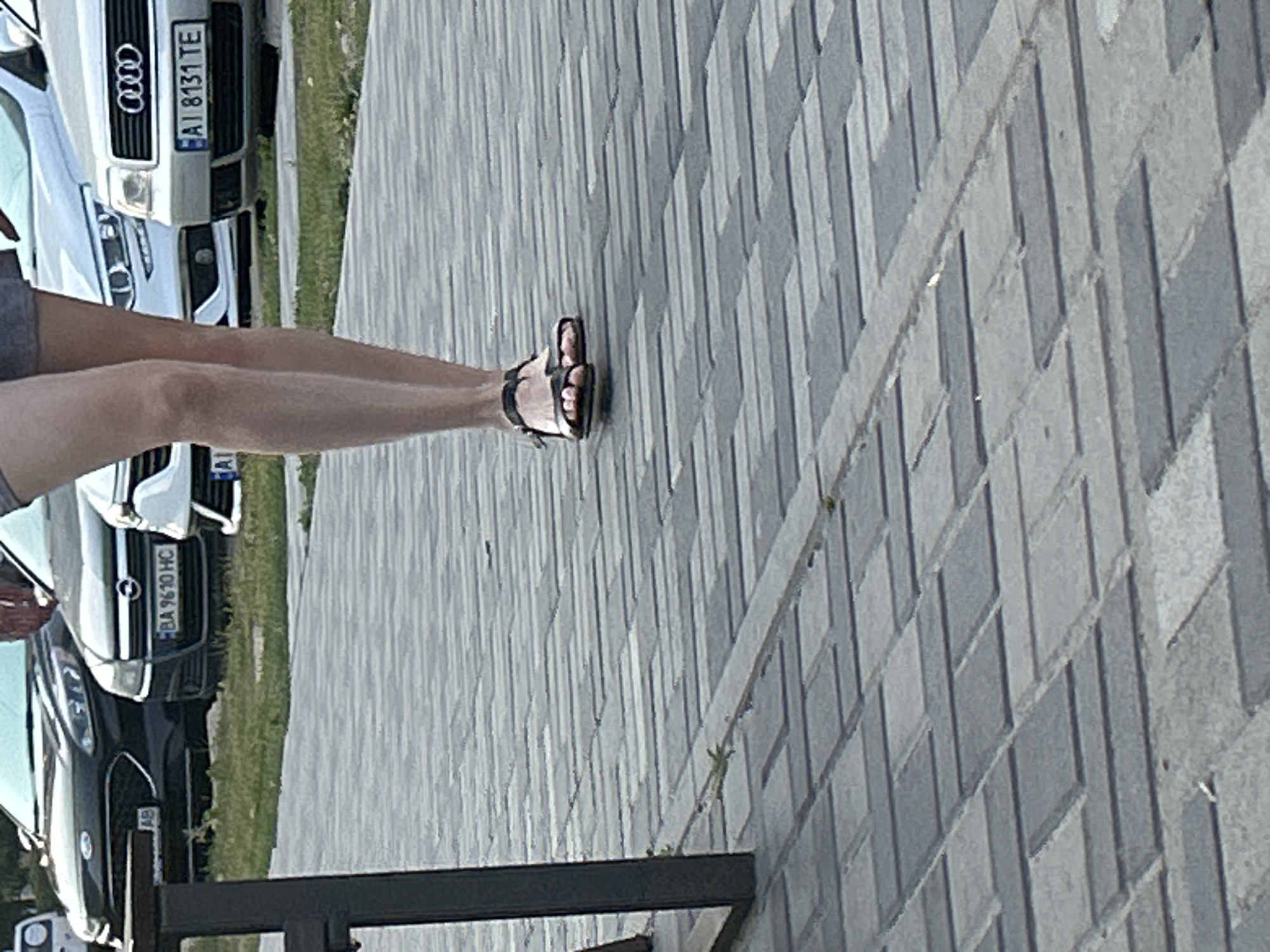 The image depicts a person's legs and feet wearing black sandals, walking on a paved sidewalk. The sidewalk is made of interlocking rectangular gray pavers arranged in a geometric pattern. In the background, several cars are visible. On the far left, the front of a dark-colored Ford Focus is partially visible, with its license plate reading "AB". To its right, a silver Audi A4 is parked, displaying the license plate "AI 8131 TE". Further to the right, another silver car is parked, and its license plate is not fully discernible. The cars are parked in front of a grassy area. The overall scene suggests an outdoor urban setting, possibly a parking lot or street in Brovary, Ukraine, given the license plate formats.