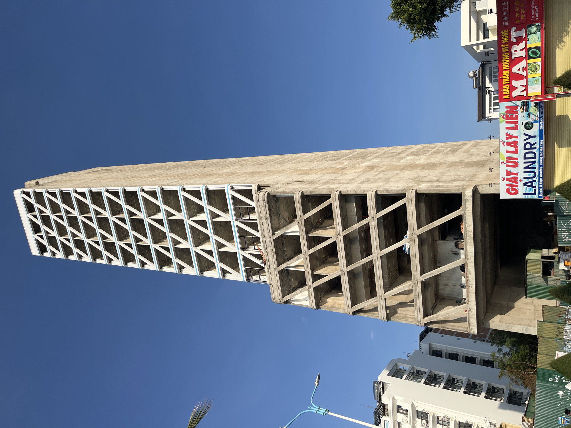 The image shows a tall, modern concrete building in Bac Nha Trang Ward, Vietnam, distinguished by its cross-lattice facade. A laundry sign, "GIẶT ỦI LẤY LIÊN LAUNDRY", is visible nearby, with a partial address indicating "49 Phạm Văn Đồng." A sign for "A BẢO TRÂM HƯƠNG MỸ NGHỆ MART" also appears in the background. The sky is clear and blue.