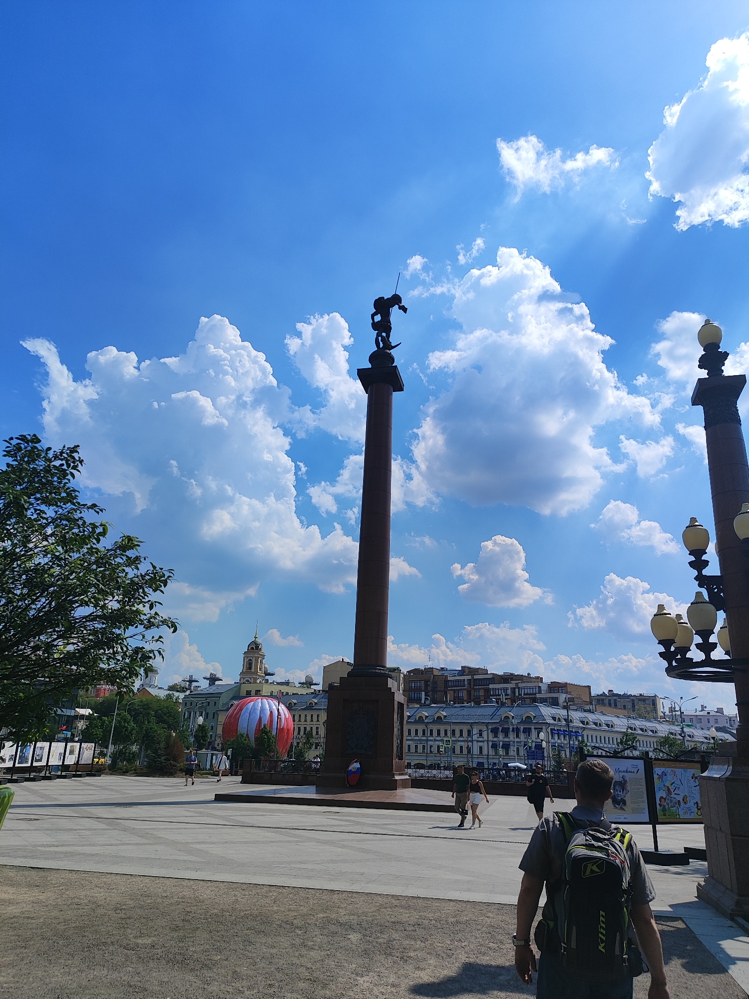 This is a low-angle, full-shot photograph taken on a sunny day in Moscow, Russia, likely at the Tverskaya Zastava Square, judging by the presence of the monument to Prince Yuri Dolgorukiy and the surrounding architecture. In the foreground, a man wearing a gray t-shirt and a black and green backpack walks away from the camera, his back to the viewer. He appears to be in motion, possibly walking towards the open plaza.
The plaza itself is paved with large square tiles and extends into the midground. A tall, reddish-brown granite monument topped with a statue of a warrior on horseback stands prominently in the center of the frame, reaching towards the bright blue sky dotted with fluffy white clouds. To the left of the monument, a large, red and white hot air balloon is visible in the distance, partially obscured by buildings.
The background features a cityscape with multi-story buildings, some with classical architecture and others more modern. A prominent church tower with a golden dome is visible among the buildings. The overall atmosphere is one of an active public space under a clear, bright sky. The sunlight casts shadows, indicating a high sun position. Several other people are scattered throughout the plaza, some walking, others appearing to be browsing exhibits on display boards. The scene depicts a typical urban public space with a mix of historical monuments, modern life, and a lively atmosphere.
