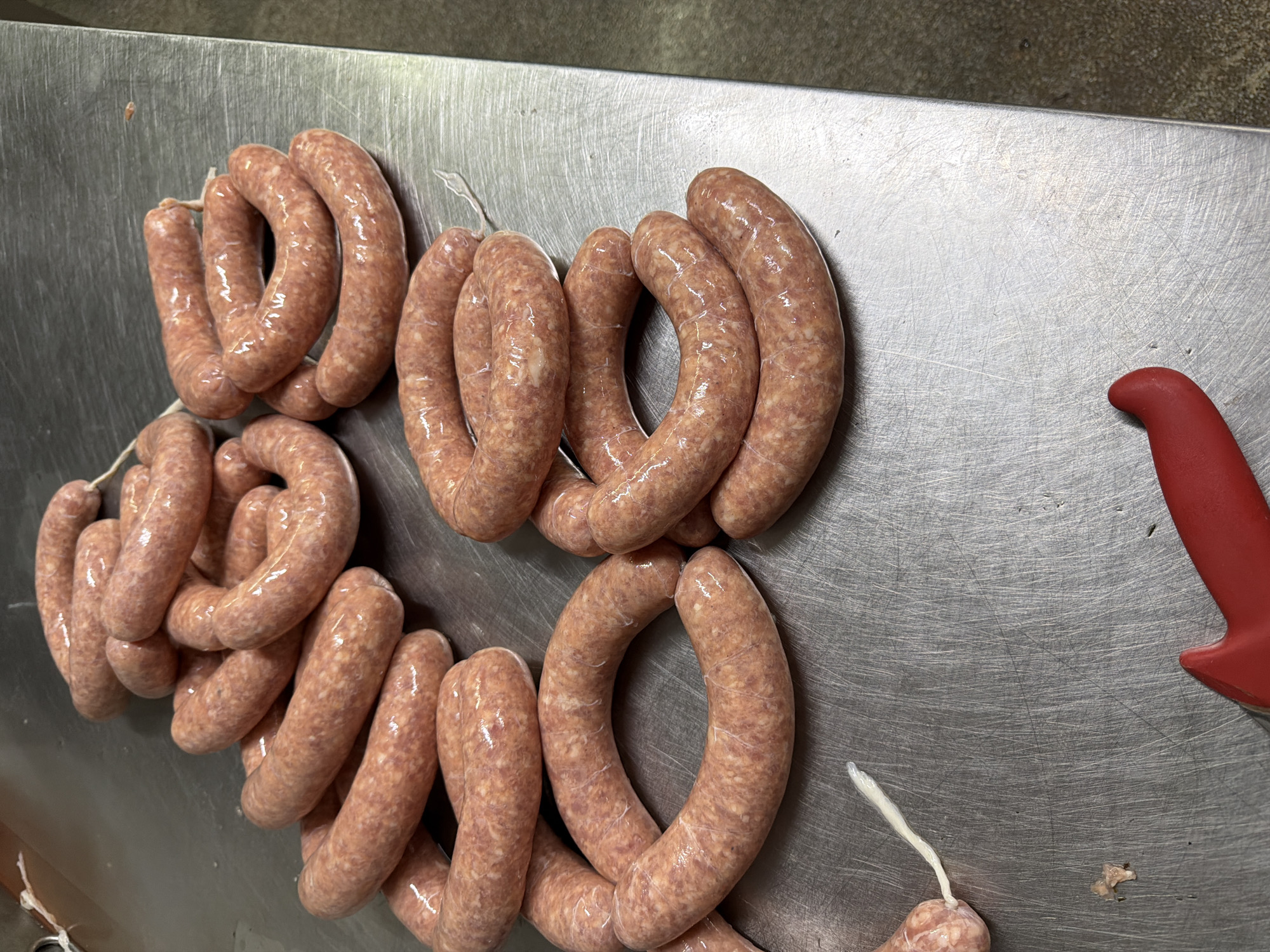 The image shows a close-up, high-angle view of several coiled raw sausages arranged on a stainless steel table. The sausages are a light pinkish-brown color and appear to be linked together with thin white twine. Some sausages are coiled into circles, while others are more loosely draped. The surface of the table is metallic and shows visible scratches and a slightly reflective sheen. In the bottom right corner of the image, the red handle of a knife is visible, suggesting that the sausages may have been prepared or are about to be processed. The lighting is bright and even, illuminating the textures and details of the sausages and the table. The scene appears to be an indoor preparation area, possibly a kitchen or a butcher's shop. There are no people visible in the image.