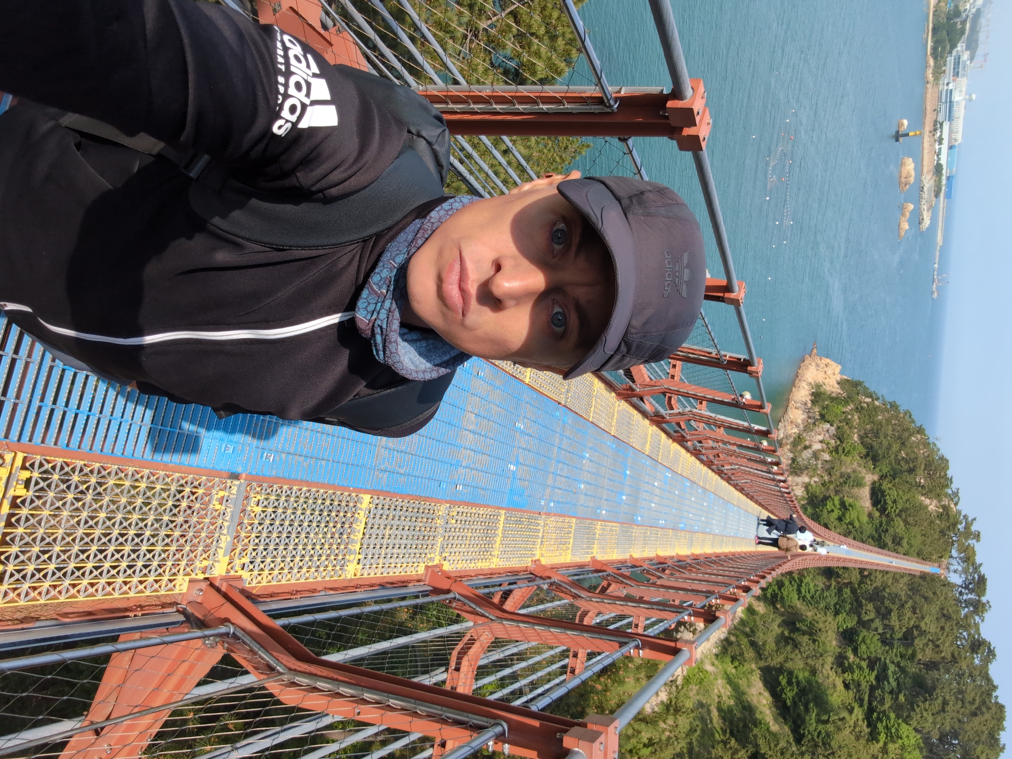 A selfie shows a person wearing a black Adidas sweatshirt, a black cap with an Adidas logo, and a blue patterned scarf on a suspension bridge. The bridge has a yellow and blue metal grate walkway and red-orange metal supports with cable railings. In the background, there is a body of water, green trees, and a strip of land in the city of Ulsan, South Korea. Two people can be seen further down the bridge.