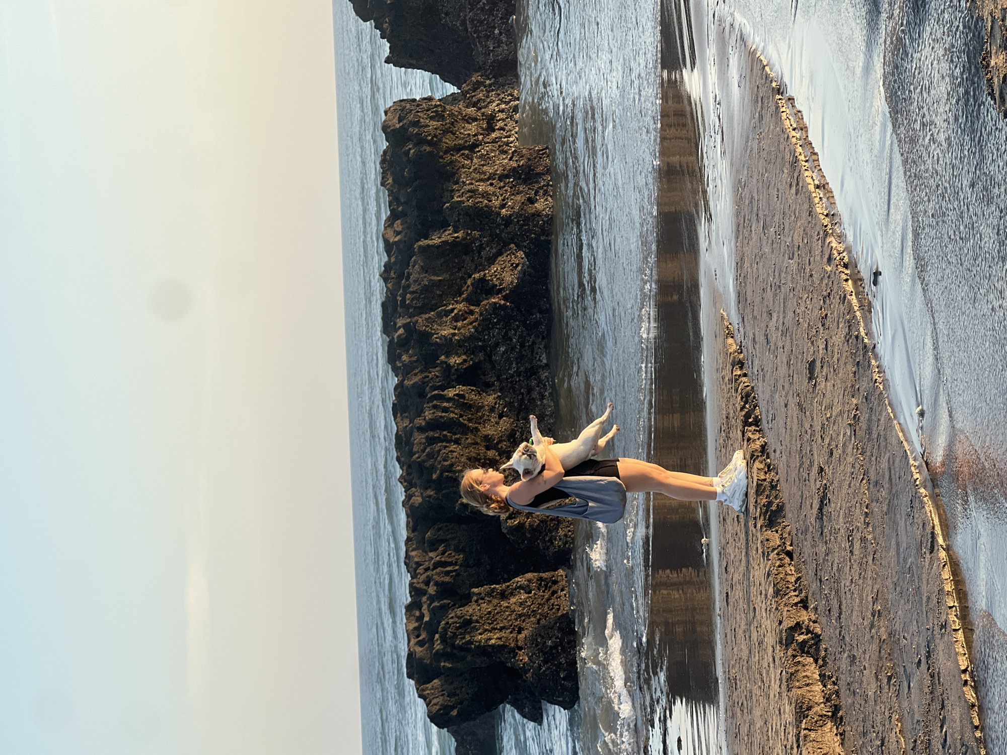 A young woman with blonde hair is holding a small white dog on a beach. She is wearing a black top, black shorts, and white sneakers. The dog is looking to the side. The woman is holding the dog in her arms and appears to be looking down at it with affection. The scene is set on a beach with dark volcanic rocks and the ocean. The water is calm, with small waves lapping at the shore. The sand is wet and reflective, indicating the tide is out or coming in. The sky is overcast, but the overall lighting suggests it is either early morning or late afternoon. The location appears to be coastal, with the volcanic rocks suggesting a volcanic island. The mood is peaceful and intimate, with the woman and her dog enjoying a moment together. There is no visible text in the image.