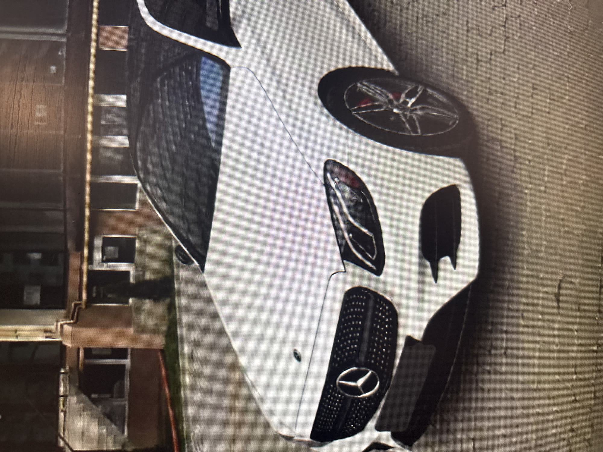 A white Mercedes-Benz E-Class sedan is parked on a paved driveway in front of a building. The car has a black roof and black grille with a silver Mercedes-Benz logo. The building appears to be a multi-story structure with brown brick and beige siding. The scene is likely located in Moscow, Russia, given the presence of a building with a distinctive architectural style common in Russian cities and the use of pavers for the driveway, a prevalent material in urban landscaping in Russia. There are no people or other vehicles visible in the image.