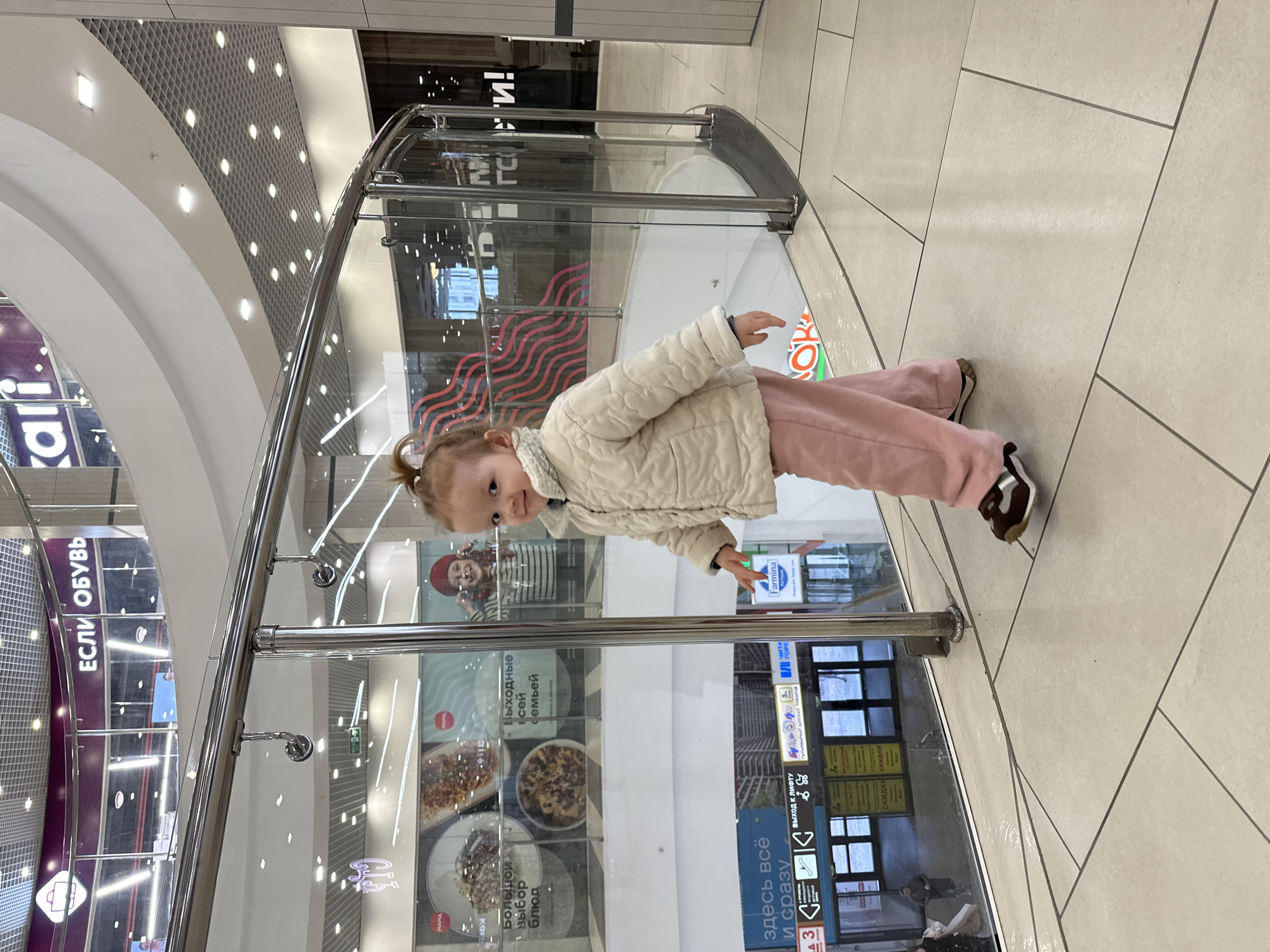 The image, originally presented sideways, depicts a young child standing in what appears to be a multi-level shopping mall. The child, a toddler with light-colored hair tied into a small bun or ponytail, looks directly at the viewer with a neutral to slightly curious expression. They are dressed in a light-colored quilted jacket, possibly white or cream, over a high-necked top, paired with pink trousers and dark shoes with white soles. Their arms are slightly outstretched.
The setting is a brightly lit, modern indoor space featuring light-colored tiled floors and prominent glass and metal railings that separate the child's level from the lower floors. The background includes curved white walls and ceilings with recessed lighting, as well as a patterned grid panel. Reflections in the glass show various shopfronts and interior structures.
Visible text on shop signs and information boards, primarily in Russian, confirms the mall environment. Examples include "ЕСЛИ ОБУВЬ" (If Shoes) and "kari." (a shoe store brand). Other signs advertise "Скидки" (Discounts) and "Выходные всей семьей" (Weekend for the whole family). Food-related signage reads "Большой Выбор блюд" (Large Choice of Dishes), indicating a restaurant or food court. Directional signs like "ВЫХОД К ЛИФТУ" (Exit to the Elevator) are also present, alongside brands such as "Farmina" and "МАМА". The scene conveys a casual outing in a bustling commercial interior.
