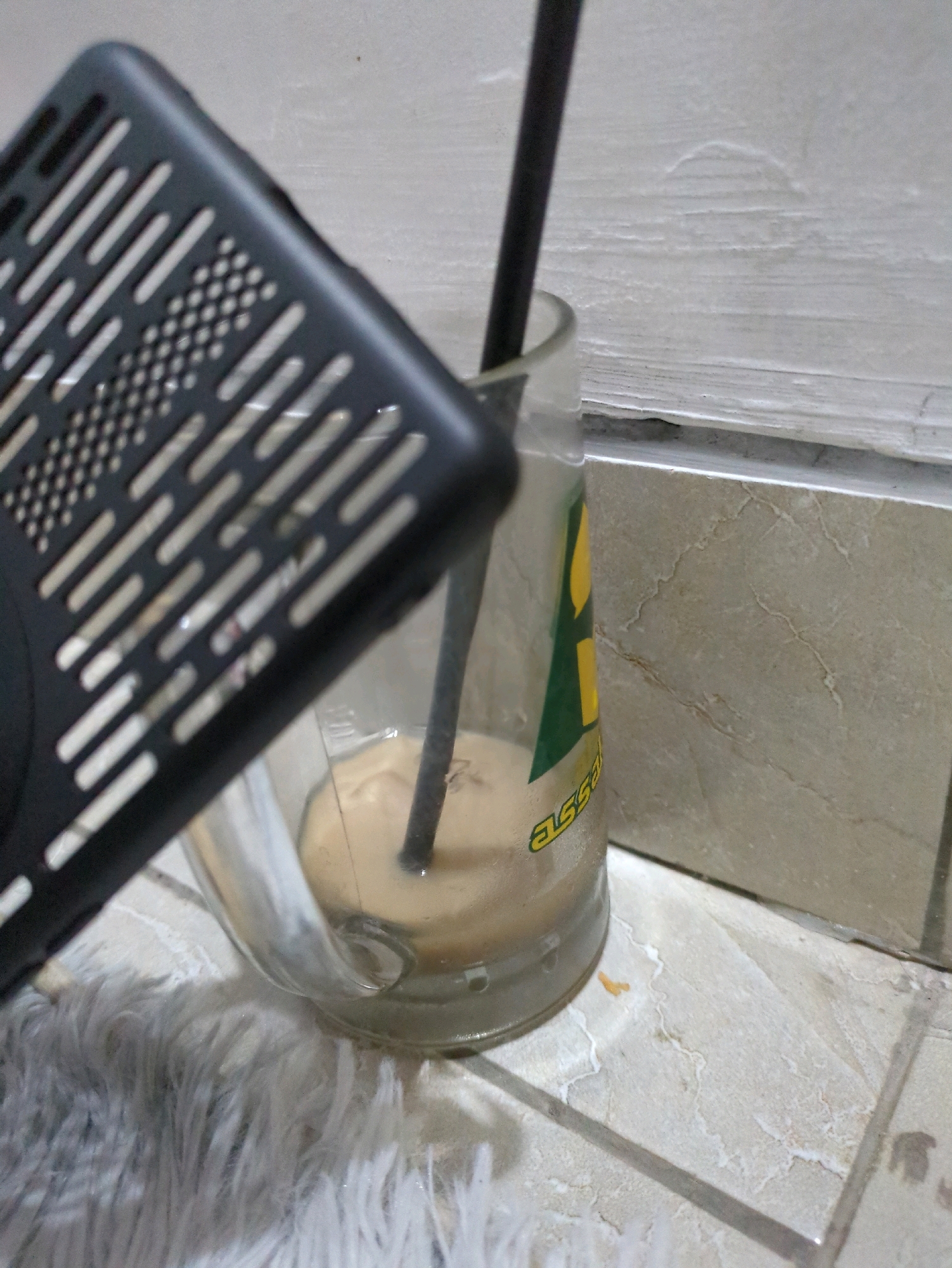 A black, perforated phone case is angled over the top of a glass tumbler containing a light brown liquid and a black straw. The tumbler is placed on a white, fluffy rug. In the background, a tiled wall with a marbled pattern is visible. The scene appears to be indoors, possibly in a home, and does not provide any specific indicators of being located in Sindon, Indonesia.