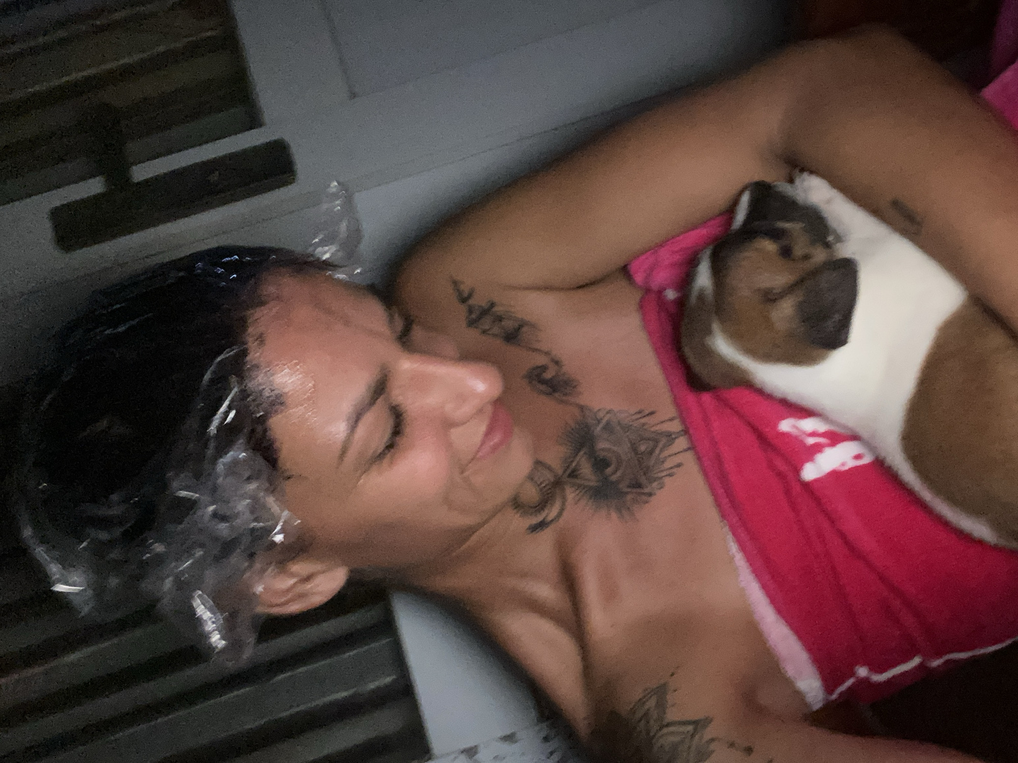 A woman with short, dark hair covered in a clear plastic shower cap is lying down, eyes closed, with a brown-and-white dog snuggled against her chest. The woman has several tattoos on her chest, neck, and arm. She is wearing a pink top. The location is identified as Mũi Né, Vietnam. A window frame is visible in the upper left corner of the frame.