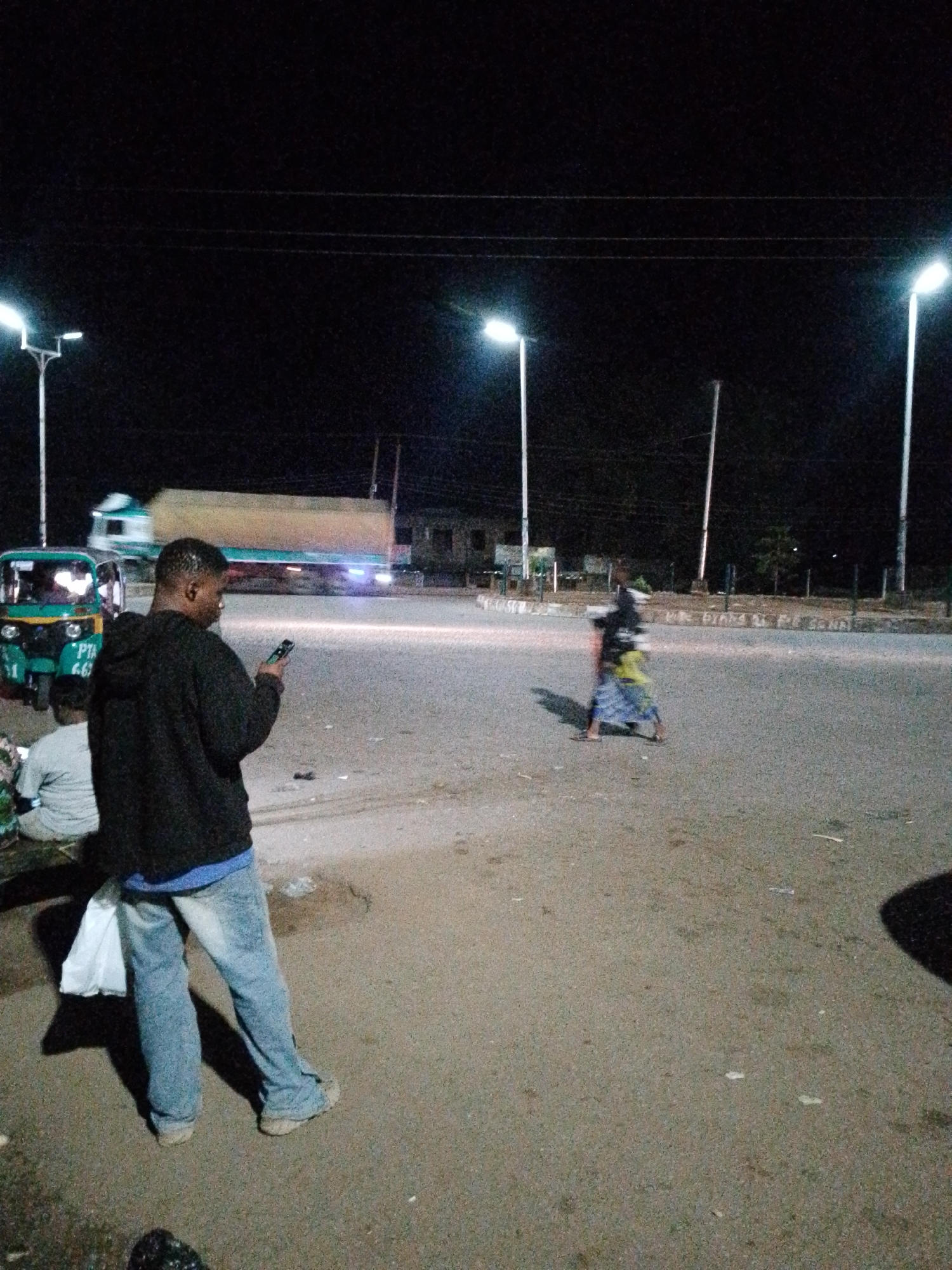 The image shows a nighttime scene at an intersection in Unknown, Unknown. Several streetlights illuminate the area, casting shadows and highlighting a truck in motion. In the foreground, a man wearing a dark hooded sweatshirt and blue jeans is looking at his phone. He is holding a white plastic bag in his left hand. To his left, another person is seated, partially obscured. In the background, a green auto-rickshaw is parked, and a blurry figure is walking in the street. The ground is paved asphalt, with scattered debris. The environment suggests a busy urban area with pedestrian and vehicular traffic.