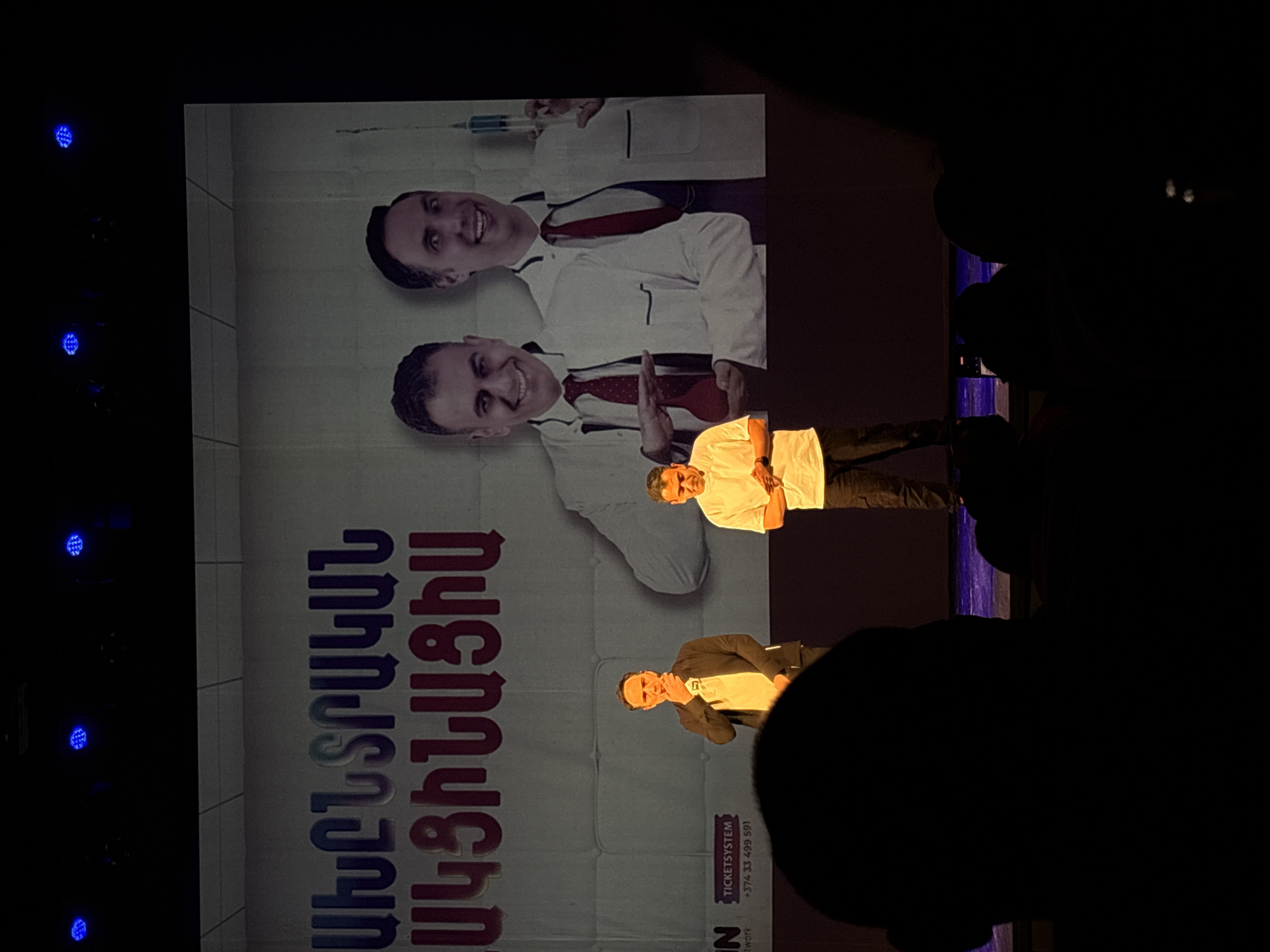 The image shows a live performance on a stage, possibly a comedy show or play. In the background is a large banner featuring two images of the same man in a doctor's coat with a syringe, above an Armenian text that reads "ՔԻԼԻՆԻԿԱԿԱՆ ՎԱԳԻՆԱԼԻՍՏ". Two men are on stage, one in a suit jacket and another in a white shirt. The performance is taking place indoors, likely in a theater or venue. There are stage lights on the left side of the banner.
It is unknown if the location is in Yerevan, Armenia, as this is not visible.