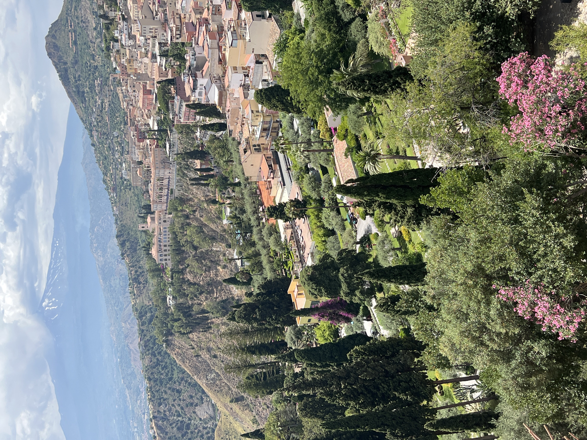 The image is a high-angle shot of Taormina, Italy, with Mount Etna visible in the background. The foreground features lush green trees and bushes, with some vibrant pink flowers. In the middle ground, there are numerous terracotta-roofed buildings nestled on a hillside, indicative of the town's architecture. A network of streets and pathways winds through the urban landscape. The background is dominated by the majestic Mount Etna, partially shrouded in clouds, with snow visible on its summit. The sky is partly cloudy, with a mix of sun and overcast conditions. There are no visible people or animals in the image. The scene depicts a scenic view of the town and its natural surroundings on a bright day.