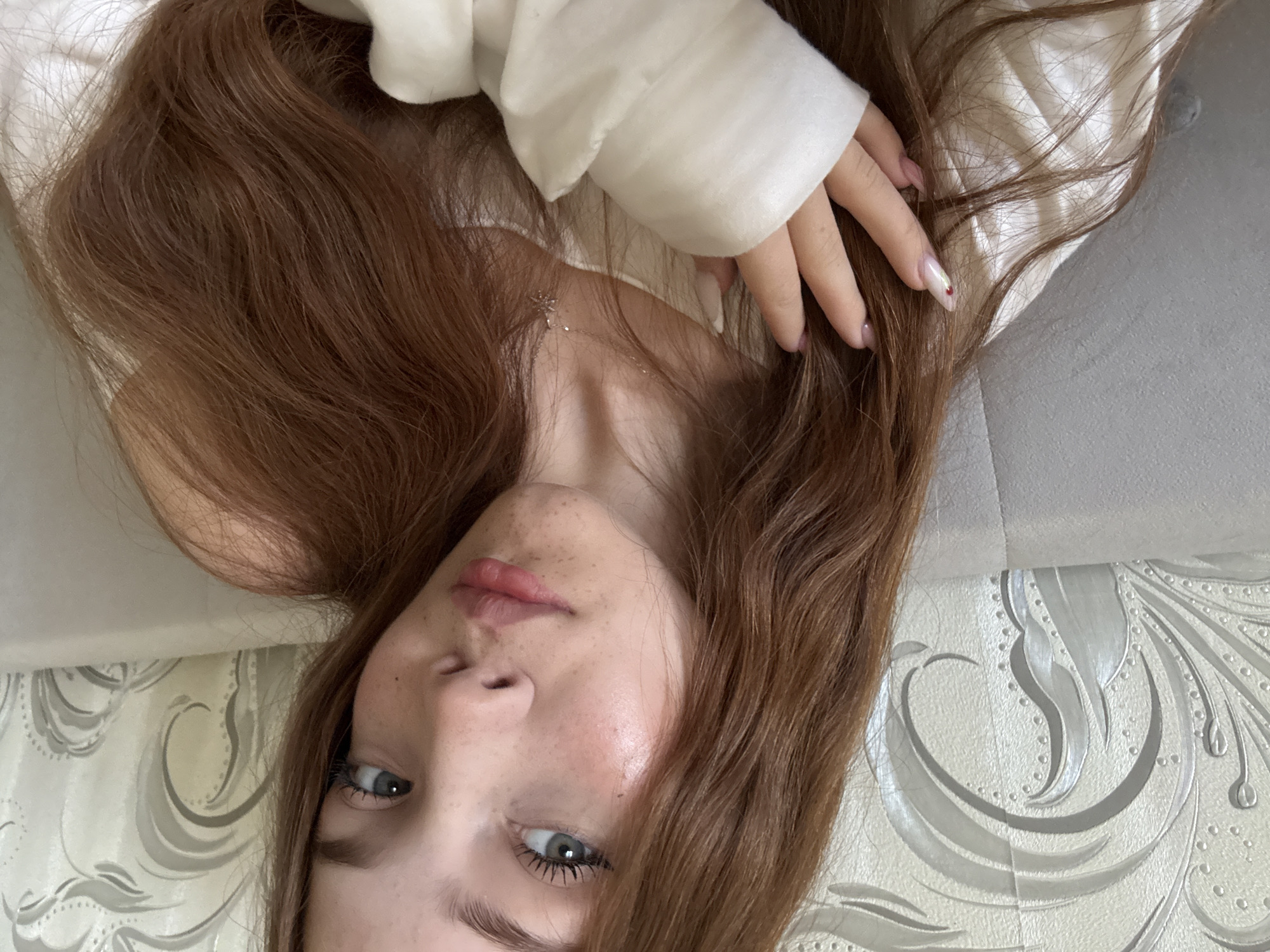 An overhead, rotated portrait features a young woman lying down indoors, likely in Naberezhnye Chelny, Russia. Her long, reddish-brown hair spreads across a light-colored surface, possibly a bed or couch. She looks directly at the camera with her left eye, which is a light grey-blue. Her face shows a scattering of freckles across her nose and cheeks, and her lips are full and slightly parted.
She is wearing a light-colored, long-sleeved garment, possibly a sweater or top. A delicate silver-colored necklace with a star-shaped pendant is visible on her neck. Her right hand, with manicured nails featuring a light pink base, white tips, and a small red dot on one, rests on her hair. The background behind her head features a light-colored patterned surface, possibly wallpaper or a decorative panel, adorned with ornate swirling designs in light grey and silver tones. The activity is a posed photograph, with the subject displaying a neutral and direct expression. No visible text is present in the image.