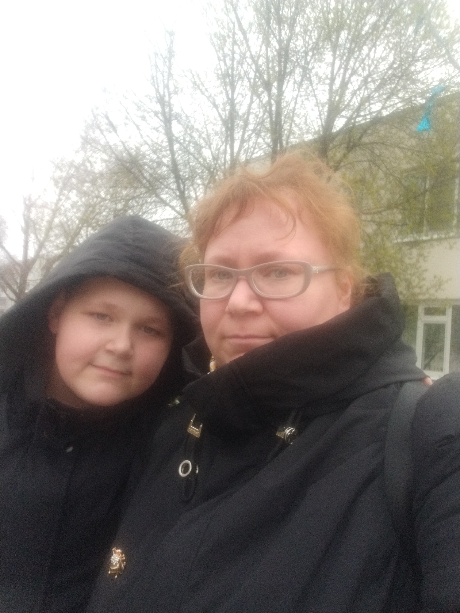 A close-up outdoor selfie captures two individuals: an adult woman and a child, likely a pre-teen, standing together. Both are wearing dark-colored, possibly black, winter or early spring jackets. The child, positioned on the left, has light skin and a subtle smile, with a hood pulled over their head. The adult woman, on the right, wears glasses with light frames and has reddish-blonde hair; her expression is neutral. A small, ornate pin, resembling a bee or beetle with decorative elements, is attached to her jacket lapel.
The background features numerous trees with bare branches or very sparse new foliage, indicating a late winter or early spring season. The sky is bright and overcast, creating soft, diffused lighting. Part of a light-colored building with dark window frames is visible behind the woman on the right, suggesting an urban or suburban environment. The individuals appear to be posing for the camera. There is no visible text in the image.