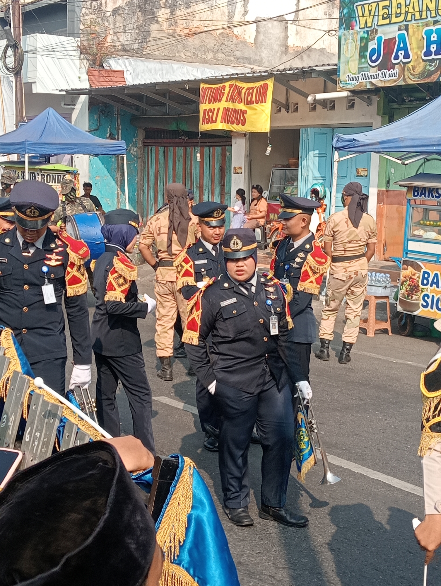 A street scene in Bintoro, Indonesia, depicts a marching band participating in a parade. Several individuals in dark blue uniforms with gold trim and epaulets are positioned on a paved road. One individual in the foreground is playing a glockenspiel. Another person, also in uniform, holds a small trumpet with a decorative banner. Behind them, individuals in tan camouflage uniforms stand at attention. In the background, there are buildings, some with faded paint and awnings, and a food cart selling bakso. Banners advertising "WEDANG JAHE" (Ginger Drink) and "ONTUNG TAPS TELUR ASLI KUDUS" (Kudus Original Egg Tarts) are visible. The lighting suggests it is daytime.