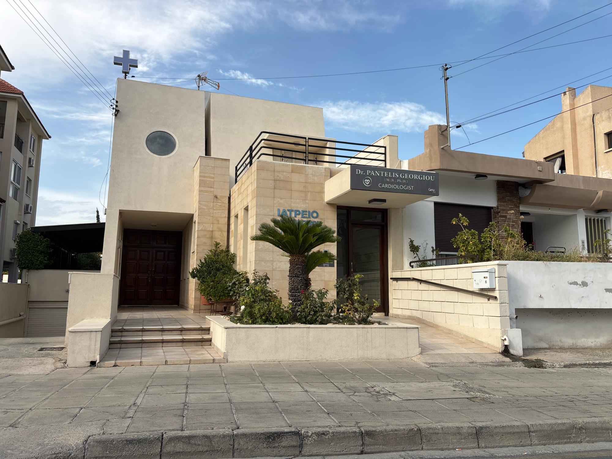 The image depicts the exterior of a medical clinic in Mesa Geitonia, Cyprus. The building is a light-colored, multi-level structure with a modern design. A prominent feature is a blue cross atop a high section of the building, suggesting a religious affiliation or a symbol incorporated into its architecture. The main entrance features large, dark brown double doors, accessible by a set of stairs. To the left of the entrance, a ramp leads up to a side entrance. A sign above a lower section of the building reads "Dr. PANTELIS GEORGIOU (M.D., Ph.D.) CARDIOLOGIST," indicating the medical specialty. Another sign on the building says "ΙΑΤΡΕΙΟ," which is Greek for "clinic." A small palm tree and surrounding landscaping are situated in a planter box in front of the clinic. The surrounding environment includes a sidewalk paved with grey square tiles and a street curb. In the background, other buildings and power lines are visible under a cloudy blue sky. The scene appears to be captured during daylight. There are no people visible in the image.
