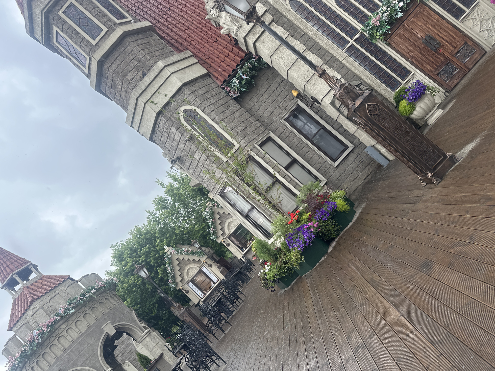 The image depicts a whimsical, fairytale-like architectural setting with elements resembling a castle or European-style buildings. The structures are made of grey stone with red tiled roofs and ornate detailing, including decorative windows, turrets, and gargoyle-like figures. Several areas are adorned with colorful flowers in planters and along the rooftops, adding a touch of vibrancy to the scene.
A wooden deck occupies the foreground, suggesting an outdoor seating area or walkway. In the mid-ground, several black wrought-iron tables and chairs are arranged, implying a cafe or restaurant setting. Lush green trees form a backdrop, interspersed with more architectural elements. One building features a large, rustic wooden door with decorative metalwork. A tall, ornate metal lamp post stands near the door.
The sky is overcast and appears to be a cloudy day. The overall mood is serene and perhaps a little mysterious, evoking a sense of fantasy or enchantment. There is no visible text in the image. Based on the provided location context, this scene is situated in Moscow, Russia.