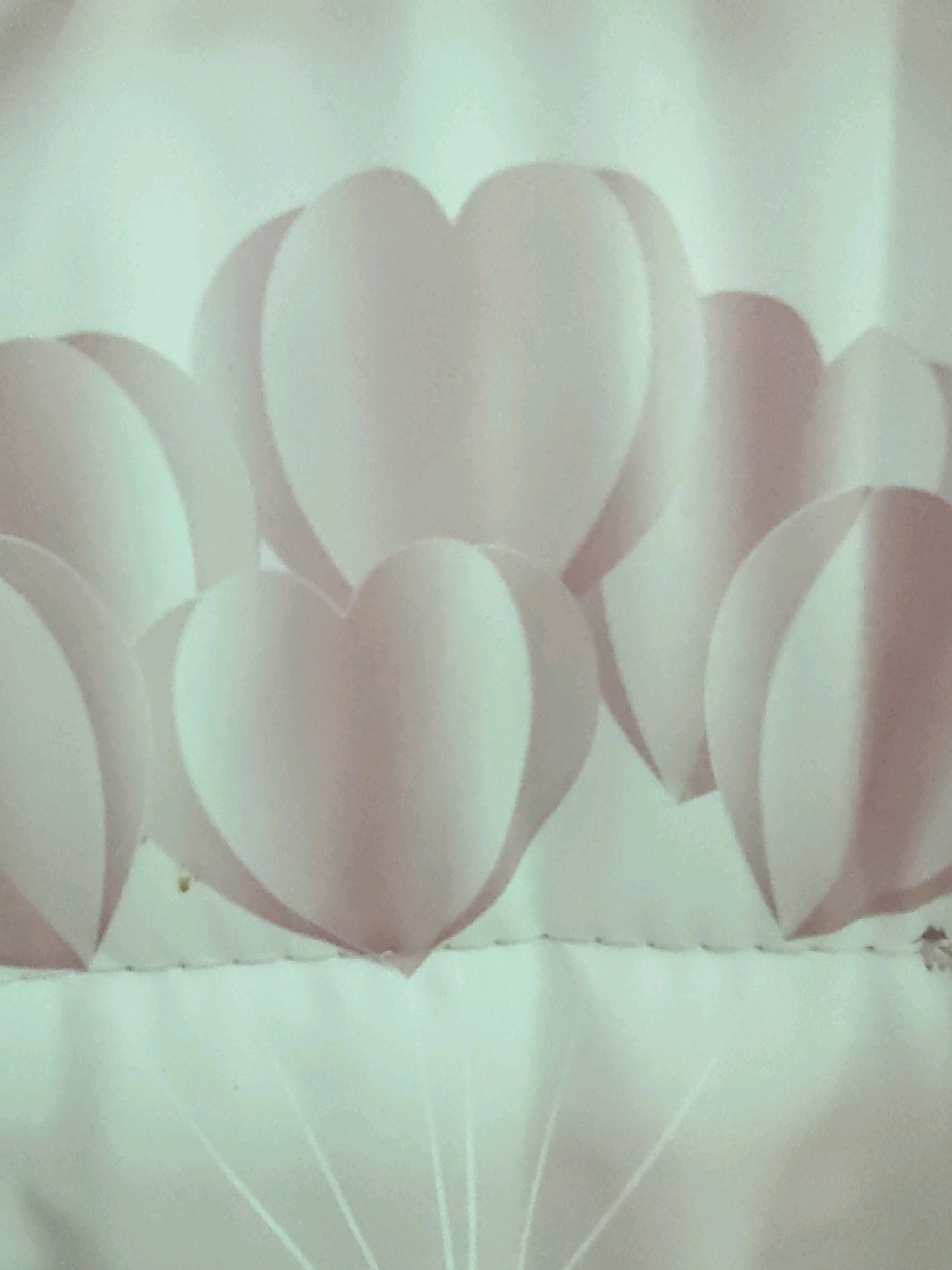 The image displays a close-up view of multiple three-dimensional paper hearts arranged in a cluster. The hearts are predominantly light pink and white, with subtle shading that gives them a rounded, inflated appearance. They are positioned in a row, overlapping and layered, creating a sense of depth. The background is a soft, out-of-focus light green or white, which helps the hearts stand out. Some of the hearts appear to be attached to thin white strings or lines extending downwards, suggesting they might be suspended or part of a decorative arrangement. The overall aesthetic is soft, delicate, and possibly festive. The image does not contain any contextual information or visual cues that would allow for its location to be identified within the city of Petroșani, Romania, or any other specific geographical location.