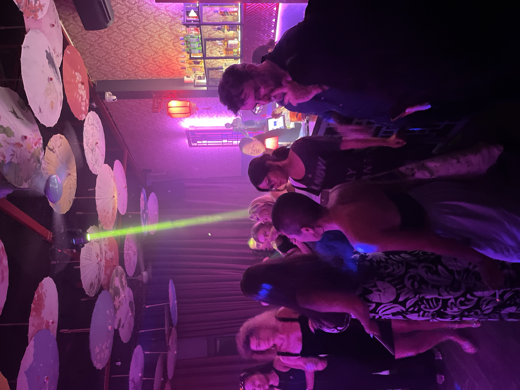 A dimly lit bar in Đà Nẵng, Vietnam is filled with patrons. A bar with bottles is visible in the background. Decorative paper umbrellas line a wall. A green laser beam shines through the room. People are gathered, socializing and drinking.