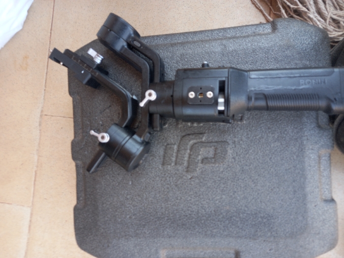 The image shows a black DJI Ronin-M camera stabilizer, lying on its side inside a black foam-lined carrying case. The stabilizer is made of black plastic and metal, with a silver knob on the left side and two silver knobs on the right side. The stabilizer is labeled “RONIN-M”. The case is made of black plastic and has a textured foam lining. The image is likely taken in Ita Oniyan, Nigeria, as the provided coordinates are located within the city. The exact location cannot be determined from the image alone. There are no people or other objects visible in the image, other than the stabilizer and its case.