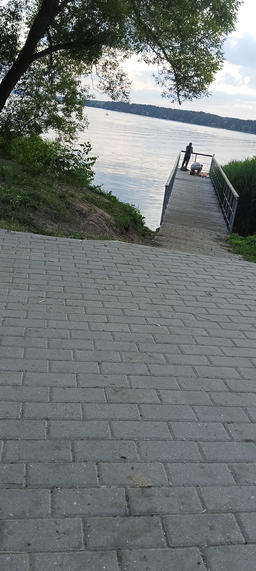 The image shows a paved walkway leading down a gentle slope towards a wooden pier on a river. The pier has metal railings and two people are visible on it. One person, dressed in dark clothing, stands at the end of the pier looking out at the water. Another person, wearing a white top and light-colored shorts, is crouched down on the pier, seemingly engaged in an activity. Trees and green foliage are present along the riverbank and overhead, with a distant tree line visible across the water. The sky is partly cloudy. This scene is likely located in the city of Konakovo, Russia, given the presence of a river and a pier typical of such a location.