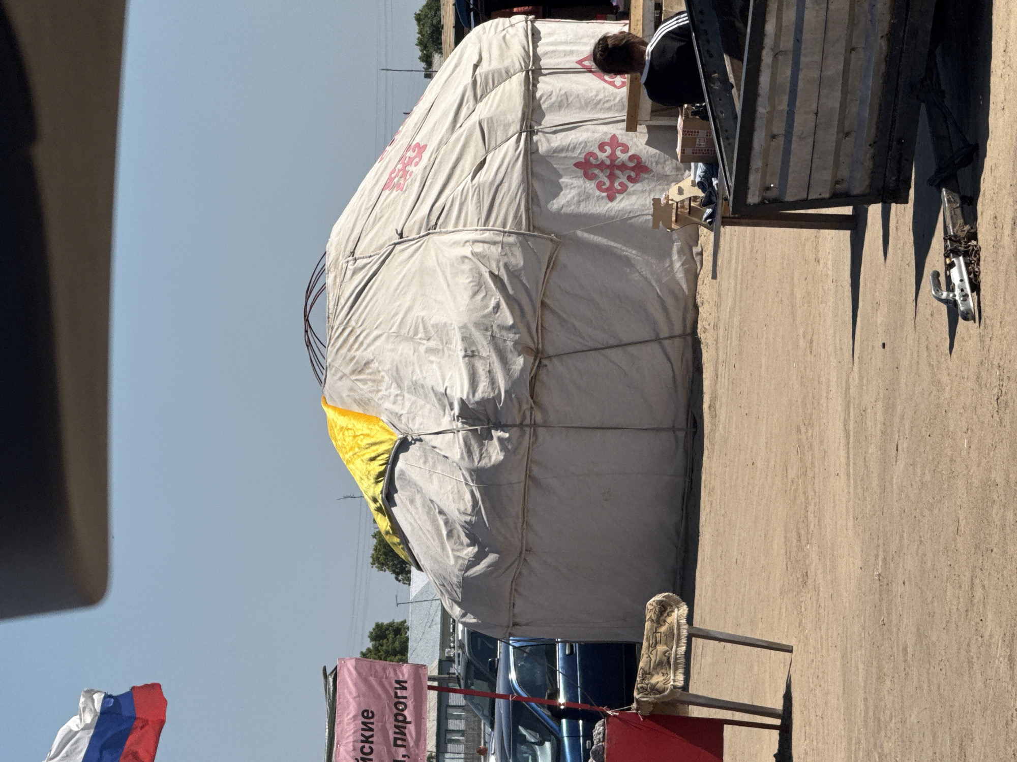 The image depicts an outdoor market scene, possibly in Бурсоль, Russia, under a clear blue sky. Dominating the center is a large, round, yurt-like structure covered in light-colored fabric, with a yellow fabric accent at the top. The yurt is decorated with pink ornamental patterns. To the left, a Russian flag, red, white, and blue, is visible against the sky. A pink banner with Cyrillic text, which includes "иские" and ", пироги", is also present. In the foreground, a metal and fabric stool with a patterned seat is positioned. Several vehicles, including a blue car, are parked behind the banner. To the right, a person wearing a black jacket with white stripes is seated, facing away from the camera. In front of them, there is a wooden table with some items on it, including boxes. A trailer with a metallic and wooden structure is prominently featured in the right foreground, with its hitch mechanism visible on the ground. The ground is sandy and appears to be unpaved. The scene suggests a temporary marketplace with goods for sale, possibly including baked goods as indicated by the banner.