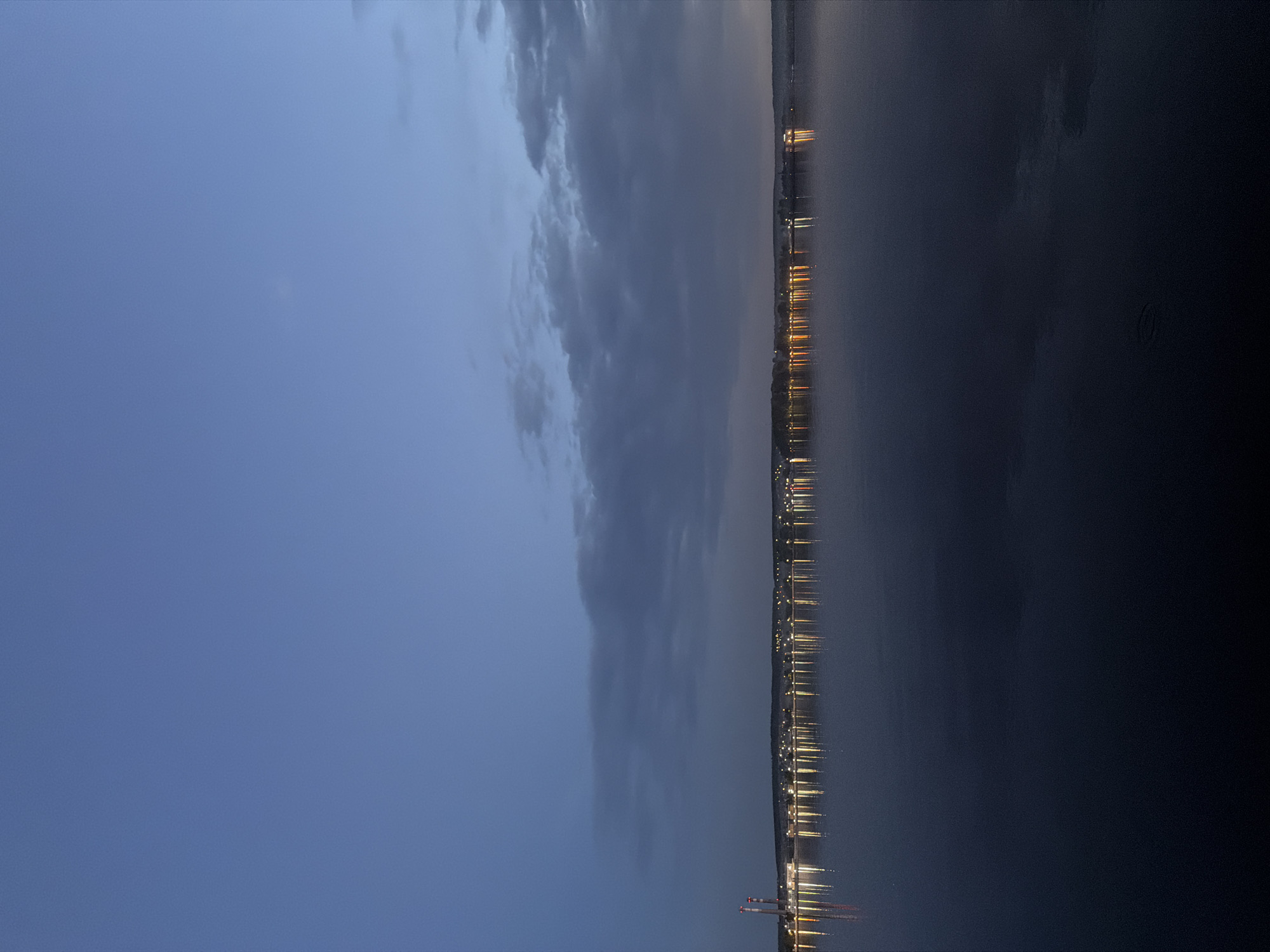 The image shows a night view of a body of water with a city skyline in the background. The sky is a deep blue, with a few clouds. The water is calm and dark, reflecting the lights of the city. The city skyline is a mix of tall buildings and smaller structures, with lights twinkling along the waterfront. There is a port facility. The location is Unknown, Unknown.