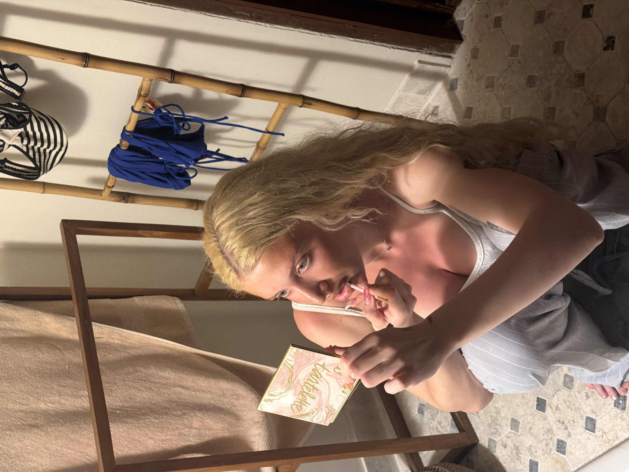 A blonde woman with her hair pulled back is captured from a high angle, kneeling or squatting indoors, intently applying makeup to her lips with a brush. She holds an open "Tartelette" makeup palette by "Tarte" (with "Sienni" also visible) in her left hand. She is dressed in a white tank top or bralette and light-colored striped bottoms. A delicate necklace with a small pendant adorns her neck, and her long nails feature French tips.
The background reveals a light-colored tiled floor with a hexagonal and square pattern. To the left, a bamboo ladder-style shelf is mounted on a wall, displaying a black and white striped bikini top and a bunched-up royal blue bikini. Shadows from the bamboo structure are cast on the wall. A wooden frame, possibly part of a bed or furniture, is visible in the foreground, adjacent to a beige towel or blanket. The lighting indicates an indoor setting, likely in the evening or under artificial light. The scene takes place in North Kuta, Indonesia.