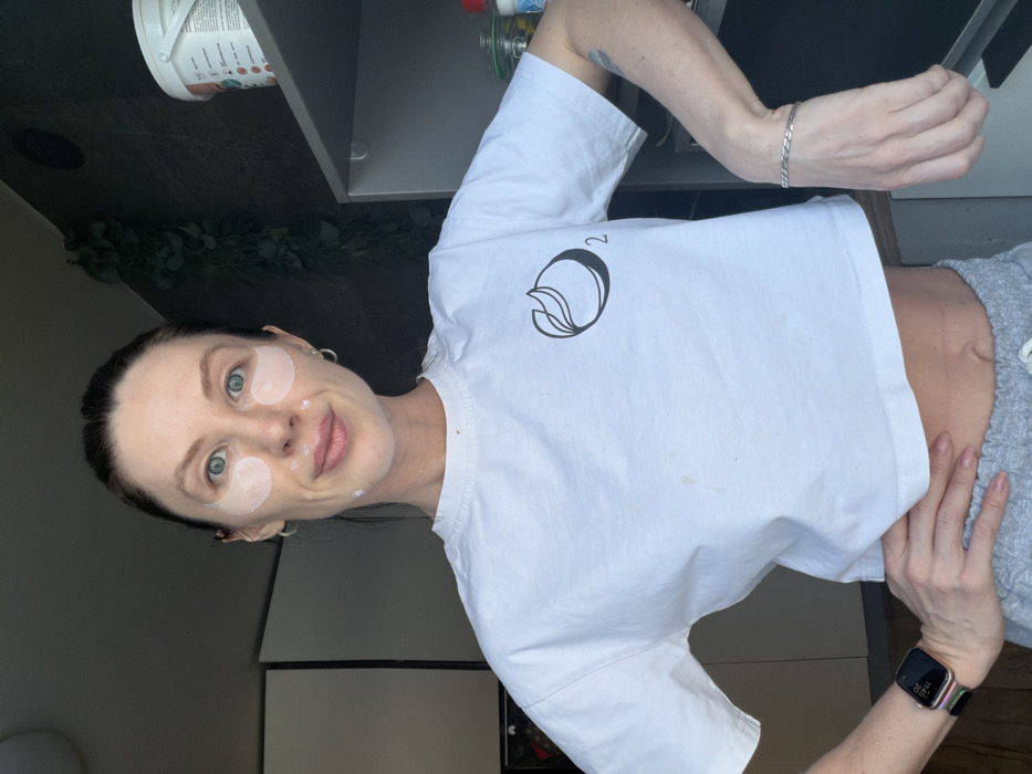 A woman with light skin, light blue eyes, and dark hair pulled back is smiling at the camera with eye patches on her face. She is wearing a white t-shirt with a circular logo on the left shoulder and grey sweatpants. A silver bracelet adorns her left wrist, and a smartwatch is on her right. A glimpse of her stomach is visible. Background elements include kitchen cabinets, a white container, and greenery. The image is rotated.