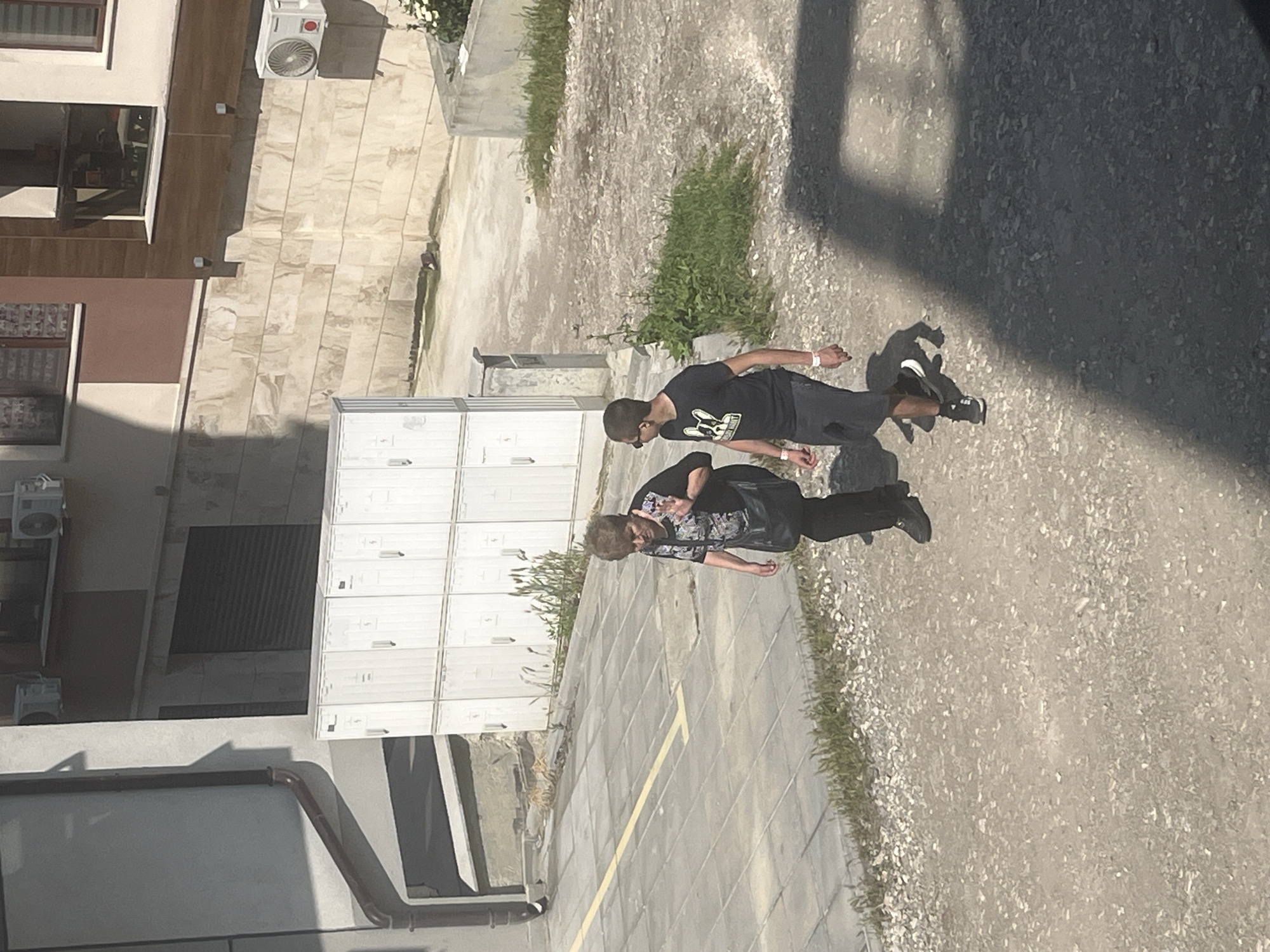 This image, presented rotated 90 degrees clockwise, captures an outdoor urban scene in Varna, Bulgaria, on a bright, sunny day. Strong shadows cast by the people and surrounding structures indicate it is daytime, likely mid-day or early afternoon.
Two individuals are walking across a varied ground surface. The person in the foreground (closer to the bottom of the original, un-rotated image) is an older woman with short, curly brown hair. She is wearing a dark, patterned short-sleeved top and dark trousers or skirt, carrying a dark shoulder bag. She looks towards the camera, extending her left hand in a peace sign, suggesting a friendly or playful interaction.
Behind her is a younger man with short dark hair, wearing sunglasses, a black t-shirt with a white graphic depicting a bulldog-like head, dark shorts or trousers, and dark sneakers with white soles. He wears a white wristband on his left wrist.
The ground consists of light-colored paved sections, possibly tiles or concrete, featuring a faint yellow line. These paved areas transition into gravelly patches with sparse green vegetation, indicative of an urban pathway or courtyard.
In the background, various building facades are visible. One structure has light-colored walls with multiple windows, some fitted with blinds. An air conditioning unit is mounted on its exterior. Another building section displays a darker, possibly terracotta-colored wall. Against one wall, a series of white, rectangular structures resembling lockers or utility boxes are stacked. Other architectural elements include a dark corrugated surface, possibly a roof or awning, and a brown gutter pipe. The overall scene depicts a casual, everyday moment in an urban environment. No clearly legible text is visible, apart from the graphic on the man's t-shirt.