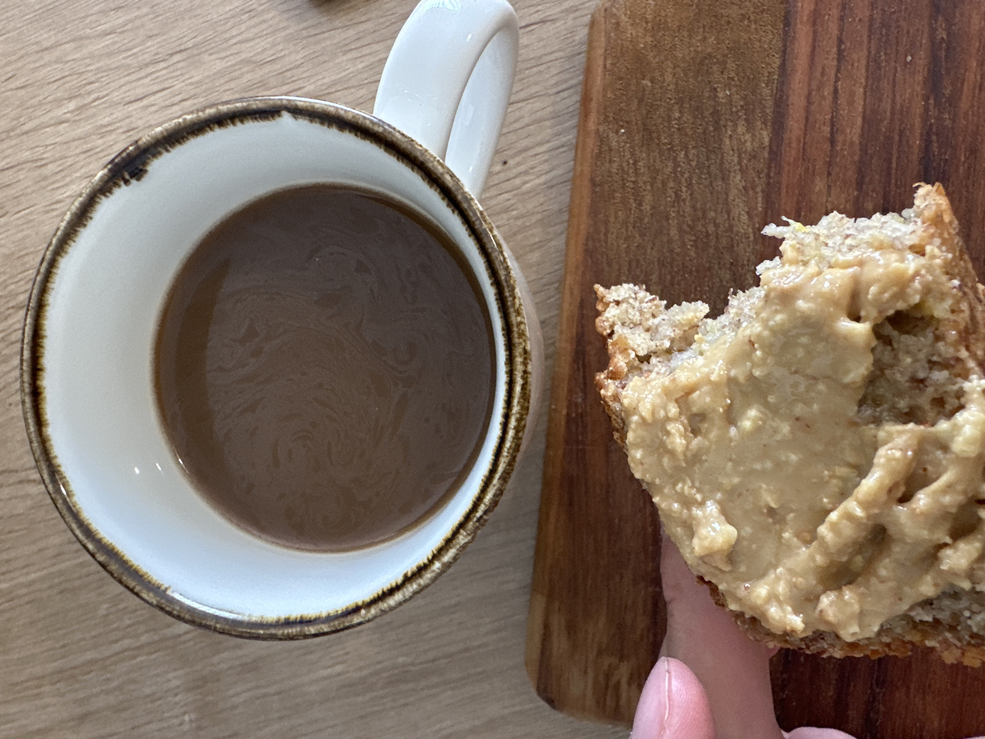 An overhead shot shows a partially eaten piece of bread smeared with a light brown spread, likely peanut butter or tahini, resting on a small, rectangular wooden board. To the left is a white ceramic mug with a brown rim and handle, filled with coffee. The items are on a light wood-grain surface. There is no indication in the photograph of the city of Fethiye, Turkey.