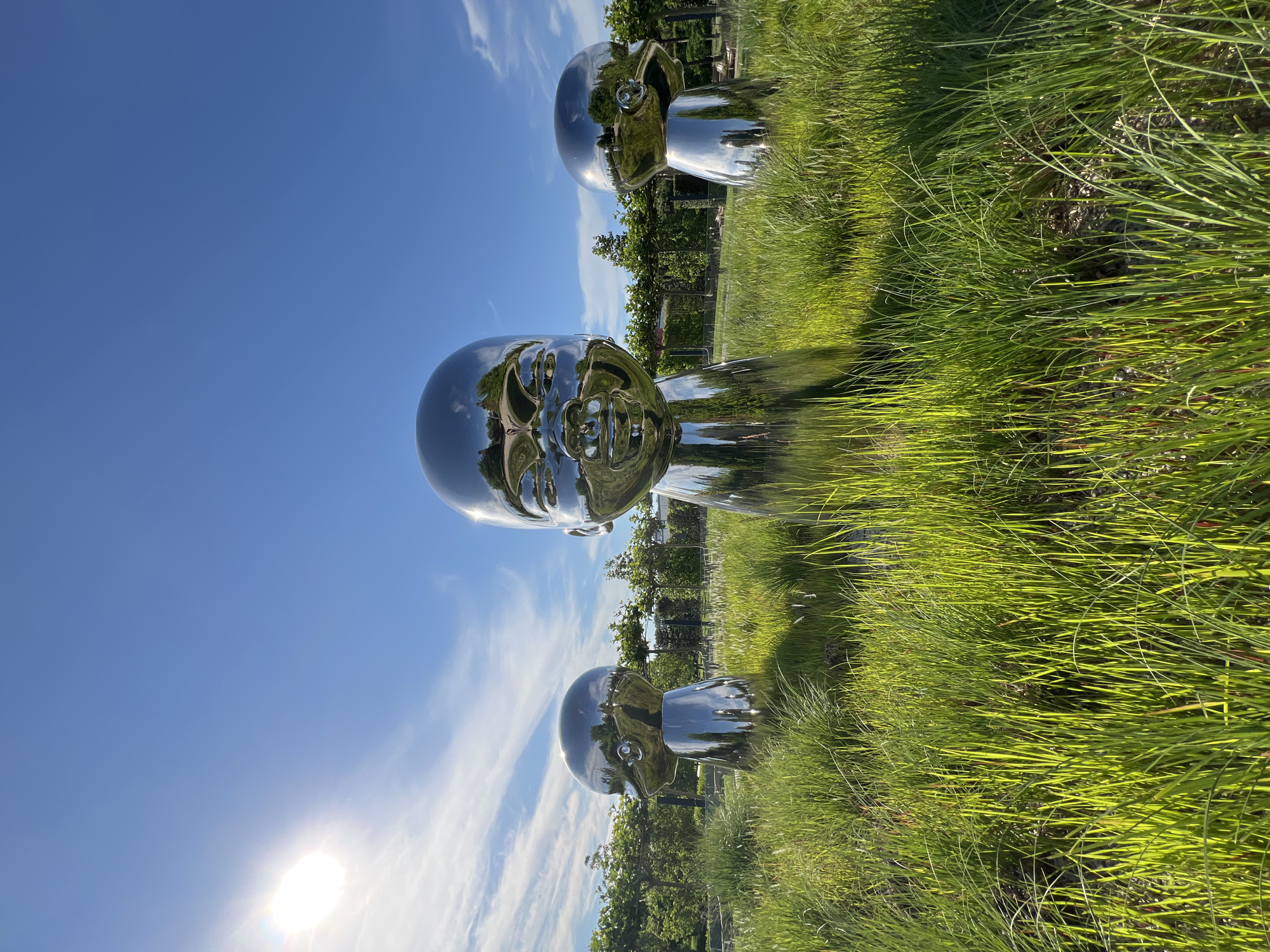 The image depicts three large, reflective, metallic sculptures of baby heads positioned in a grassy outdoor area. The sculpture in the center is the largest and most prominent, with its face turned slightly towards the viewer. Two smaller sculptures are visible to the left and right of the central one, also appearing to be baby heads. The sculptures are highly polished and reflect the surroundings, including the sky, sun, trees, and greenery.
The environment consists of tall, lush green grass in the foreground, with more manicured landscaping, trees, and some structures visible in the background. The sky is a bright blue with scattered white clouds. The sun is visible in the upper left corner of the frame, casting a bright glare and illuminating the scene.
The location of this artwork is in Krasnodar, Russia, specifically at the "The Universe of the Future" museum complex. The sculptures are part of the "New Children" exhibition by artist Li Chen.