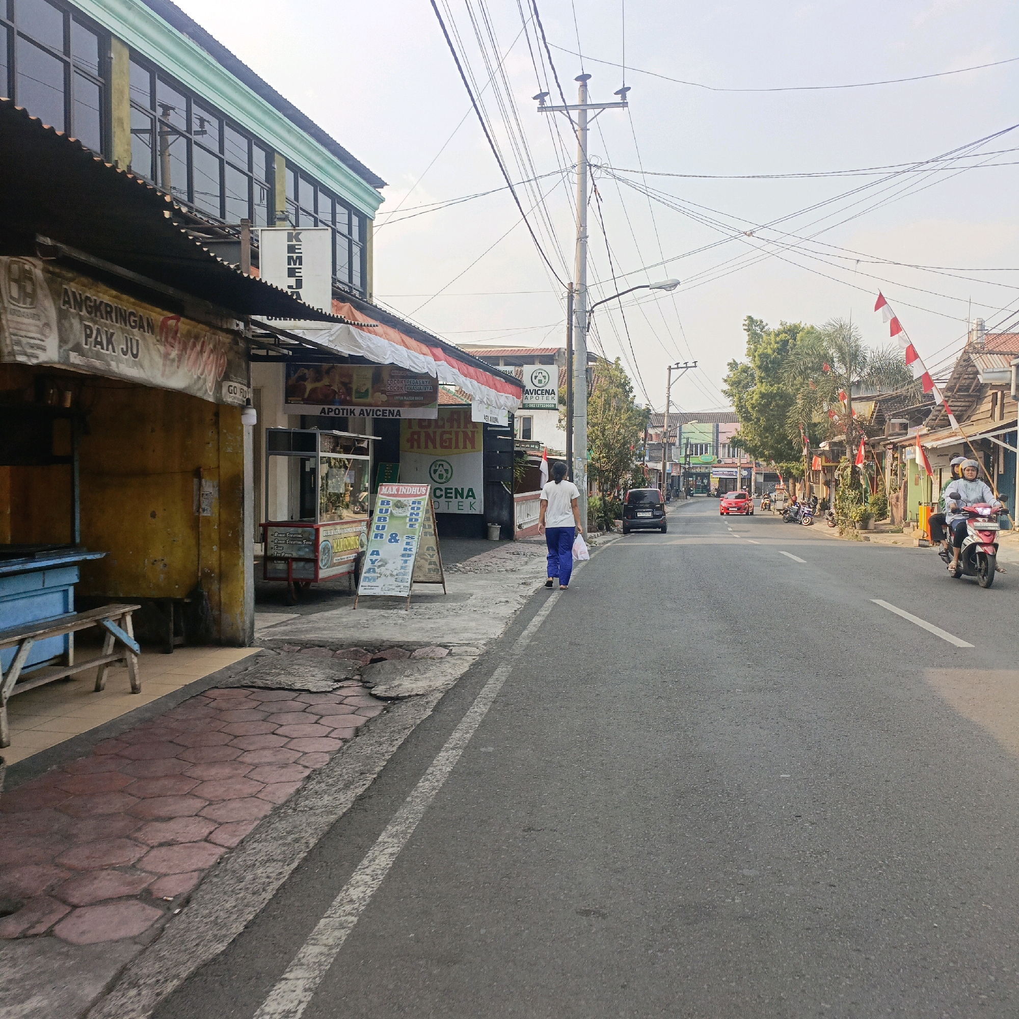 This is a street-level, eye-level photograph taken in Salatiga, Indonesia. The scene depicts a street with a sidewalk on the left and the road occupying the right side of the frame. On the sidewalk, there are several businesses, including a yellow-colored establishment with a sign that reads "ANGKRINGAN PAK JU". Next to it, there are other shops with awnings and signs, one of which says "APOTIK AVICENA". A woman in a white shirt and blue pants is walking away from the camera on the sidewalk. Further down the street, on the road, a red car and a black SUV are visible, along with several motorcycles. On the right side of the road, there are more shops and buildings, some decorated with red and white Indonesian flags, suggesting a celebration or national holiday. The sky is overcast and a power pole with numerous wires is prominent in the middle of the frame. The road surface is asphalt, and the sidewalk is paved with hexagonal tiles.