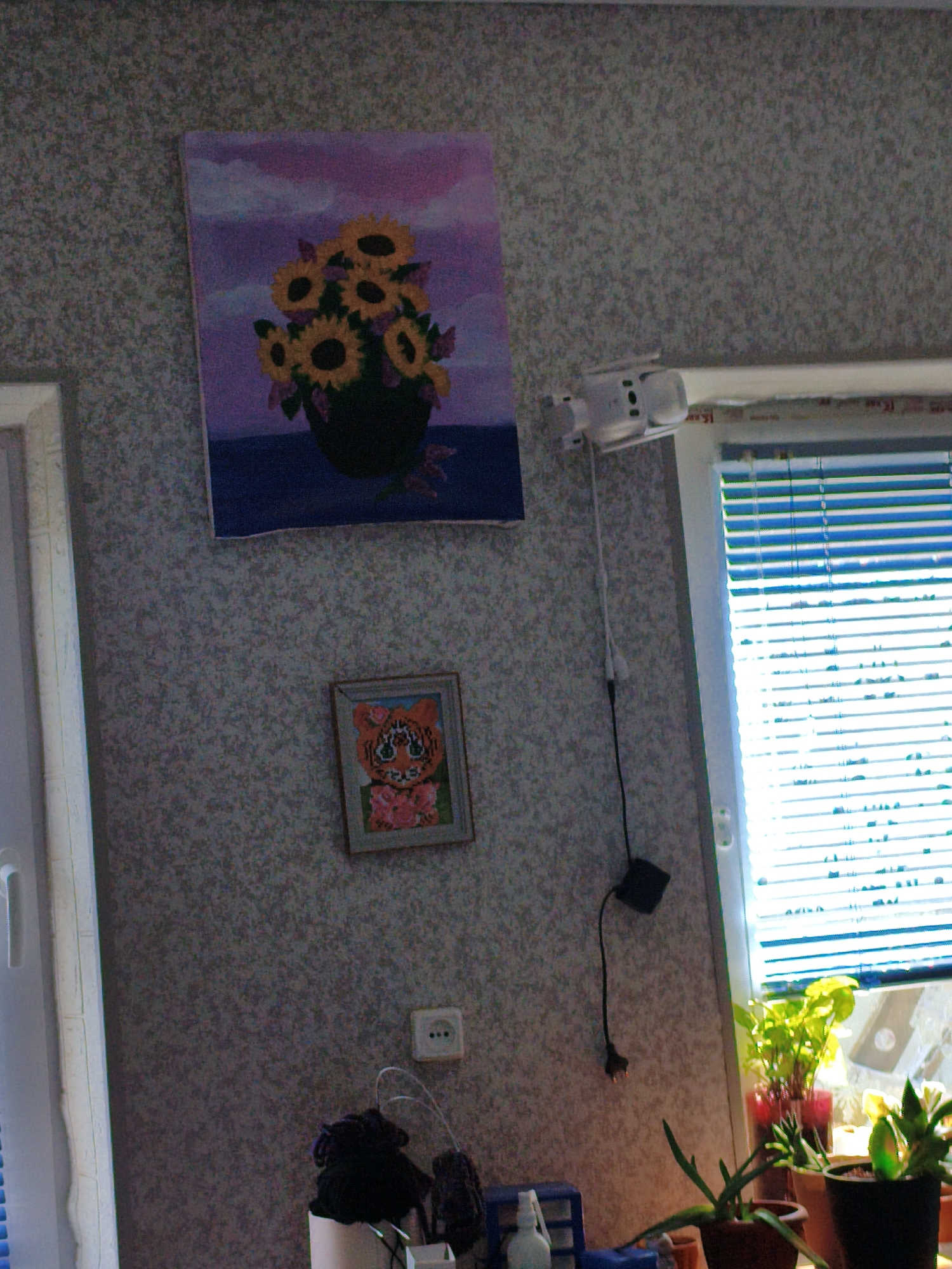 This is a slightly angled, eye-level photograph of a domestic interior. The scene depicts a wall with textured wallpaper, a window with blue blinds, and various decorative and functional items.
On the wall, there are two framed artworks. The larger one, positioned higher up, is a painting of sunflowers in a dark pot against a purple and pink sky. Below this, a smaller framed piece features a stitched image of a tiger's face, adorned with pink flowers. To the right of the tiger artwork, a white electrical outlet with three dots is visible.
A white security camera is mounted on the wall above the sunflower painting, with a black cable running down from it. Near the window, several potted plants are placed on a windowsill, with the sunlight filtering through the blinds. In the lower section of the image, a few objects are visible on a surface: a dark, possibly knitted, item and a white spray bottle.
The overall impression is of a lived-in room, likely a living space or bedroom, in daylight. The presence of houseplants and artwork suggests a personal touch. The window blinds indicate a desire for privacy or light control. The location context is Orenburg, Russia, but no specific landmarks are visible to confirm this.