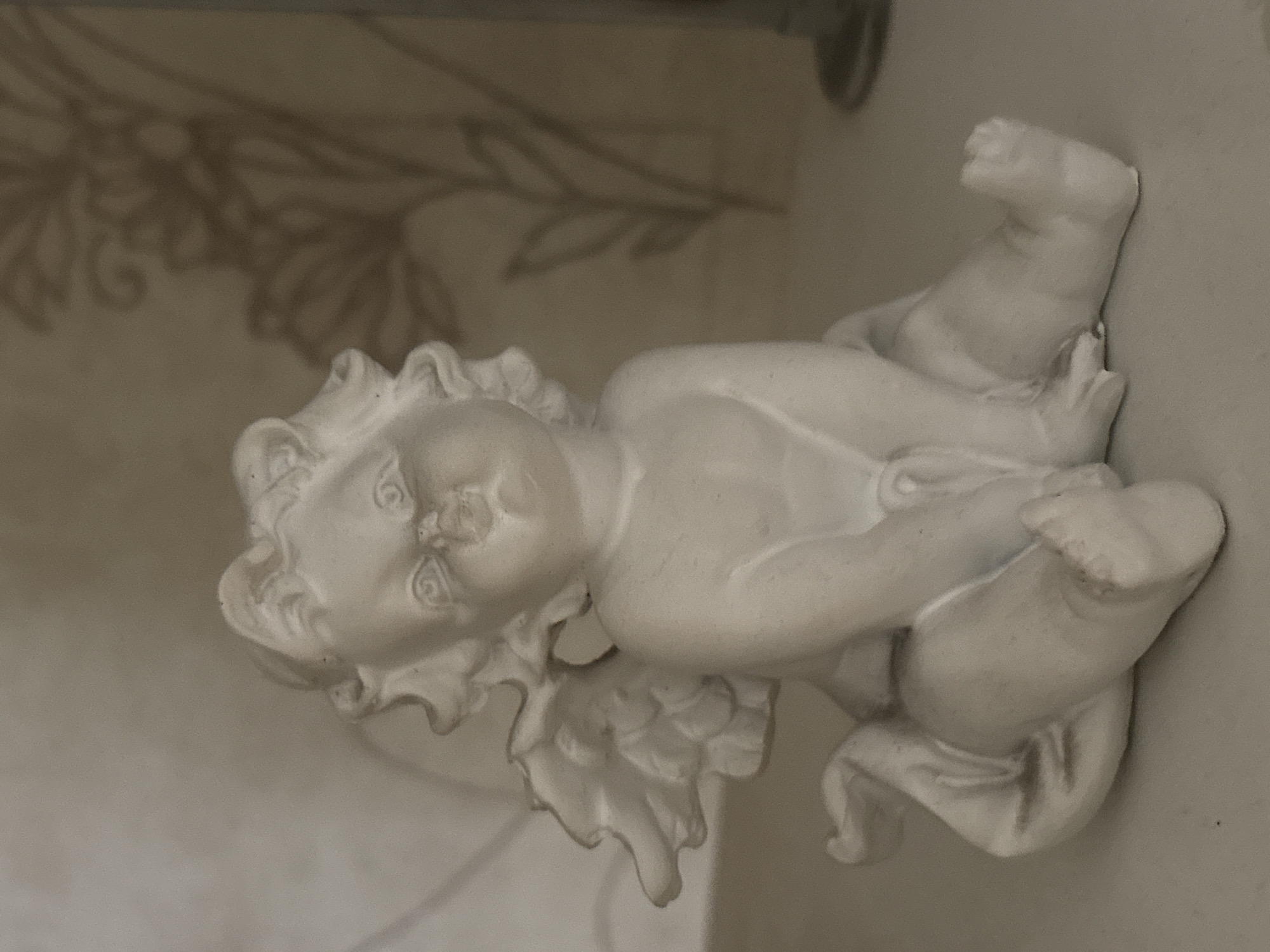 The image displays a close-up, eye-level, indoor shot of a white, plaster cherub figurine. The figurine is depicted in a seated position, with its legs crossed and its arms resting on its knees. It has small, detailed wings folded against its back and curly hair. The background is softly lit and shows a blurred floral pattern on what appears to be wallpaper. The surface on which the figurine rests is a pale, flat surface. The image quality is slightly grainy. No people, actions, or interactions are visible, and the location of Tikhiye Zori, Russia, is not depicted or suggested by the visual information within the image.