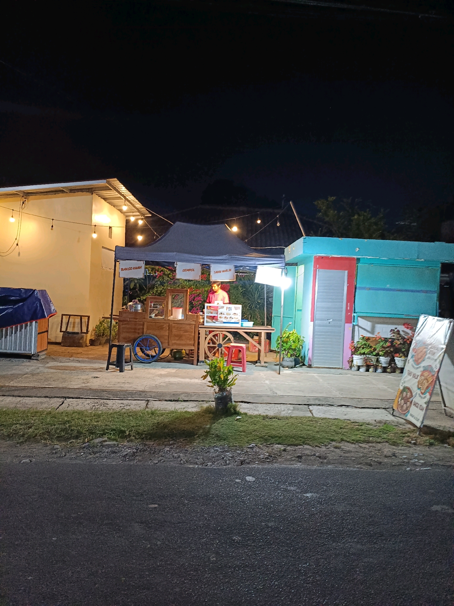 A night scene in Maguwoharjo, Indonesia, features a food stall with a person standing behind a wooden cart filled with food items. The stall is illuminated by string lights and a bright lamp. To the left, a building with a covered structure is visible. To the right, a blue and red kiosk stands with potted plants in front of it. A white easel displaying a menu is also present. The foreground shows a paved road with a grassy strip and a sidewalk. The overall atmosphere is that of a typical street food vendor operating after dark.