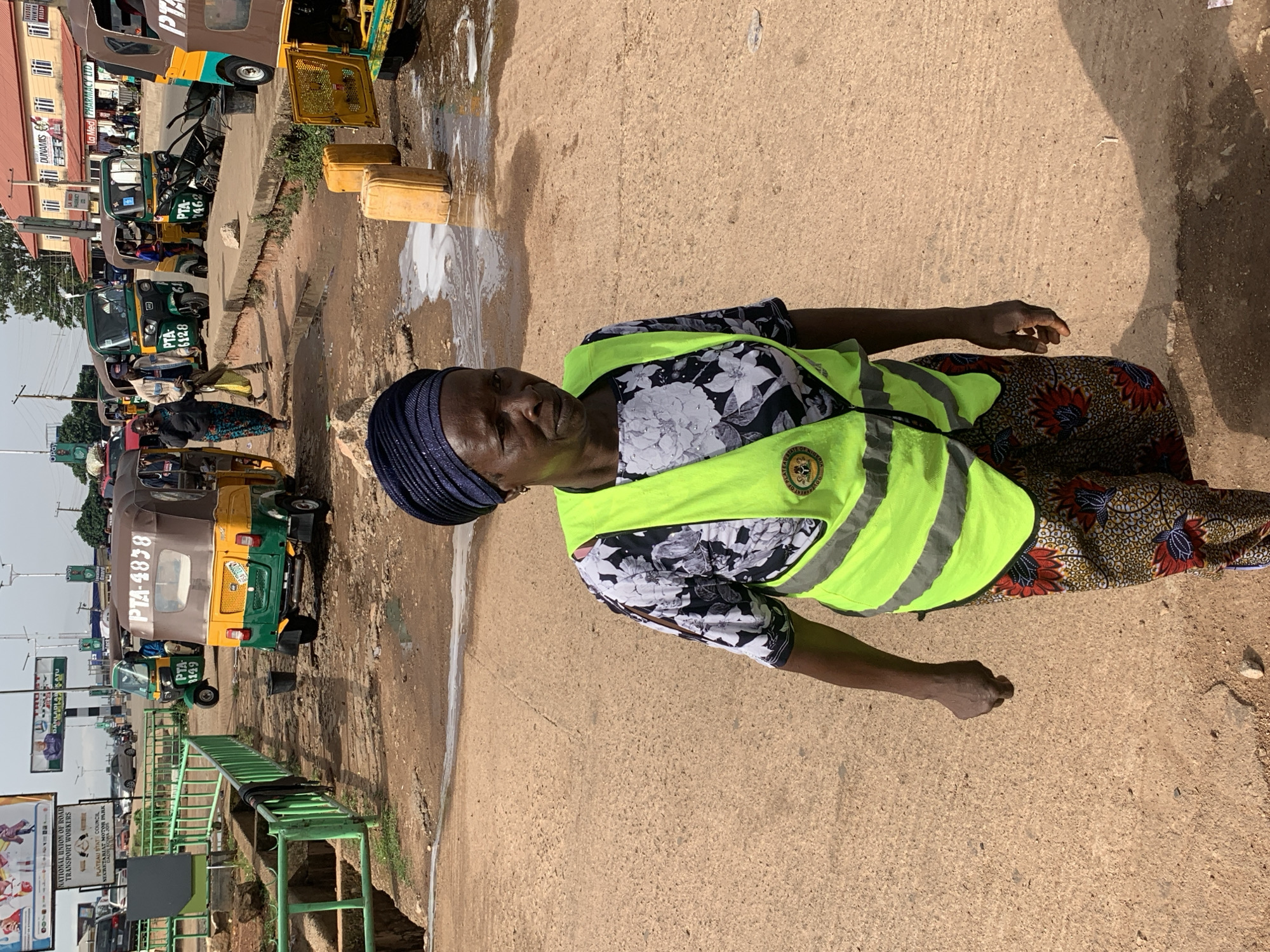 The image captures a street scene in Gura Top, Nigeria, focusing on a woman wearing a bright yellow-green safety vest over a floral print shirt and patterned trousers. She is looking to her left, with a serious expression. Her head is covered by a dark blue headscarf. In the background, several auto-rickshaws, commonly known as Keke Napeps, are parked or in motion. One auto-rickshaw in the foreground has the license plate "PTA-4828". The ground is a mix of packed dirt and some patchy concrete or asphalt, with visible puddles of water and some discarded yellow containers. The overall environment suggests a busy, possibly commercial or transportation hub within the city. Other people are visible in the background, some near the auto-rickshaws, and a poster on a pole behind them. The lighting indicates it is daytime.