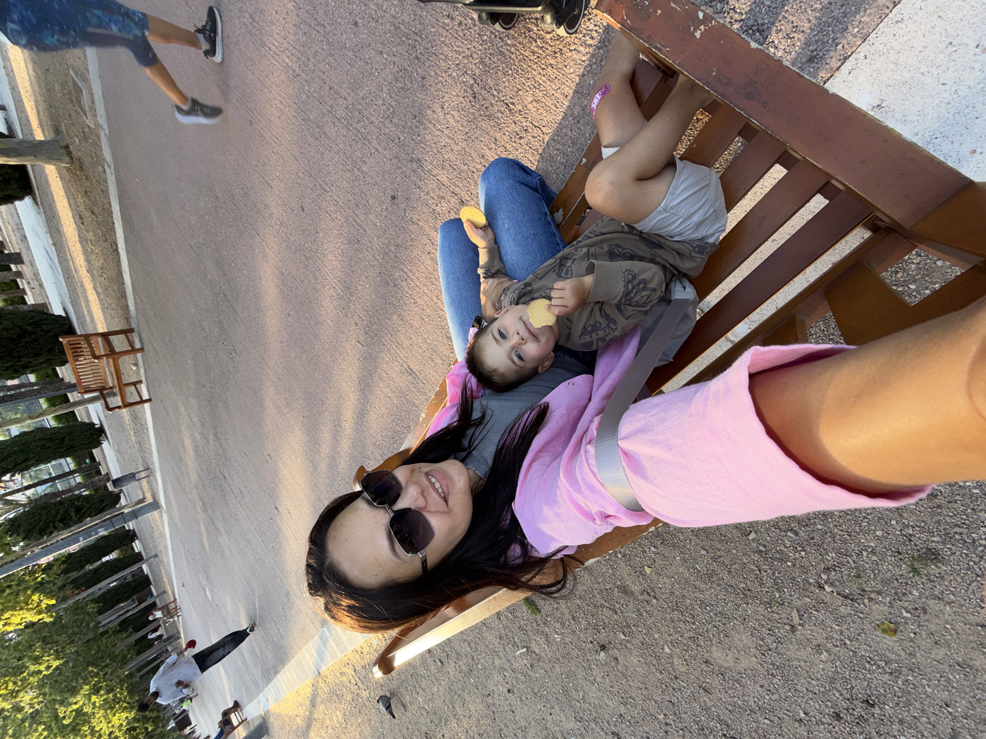 A woman is taking a selfie on a park bench with a young boy. She is wearing sunglasses and a pink jacket. The boy is holding a cookie. The park is paved with sand and gravel, and lined with trees. In the background, a man in a white shirt and dark pants is walking. Another person is running on the paved path.