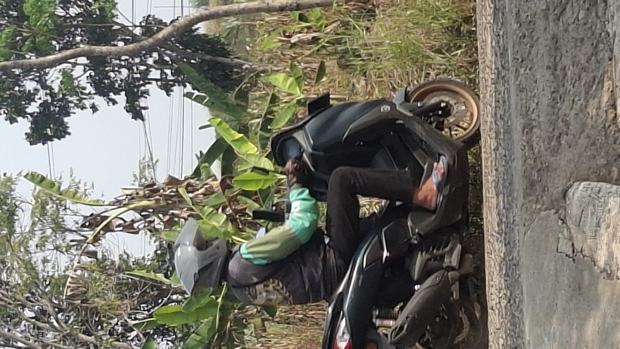 A person wearing a dark helmet and a dark vest over a teal long-sleeved shirt rides a dark-colored scooter. The person is wearing black pants and blue and white flip-flops. The scooter has a dark body with a gold-colored rim on the rear wheel. The scene is outdoors, with lush green banana trees and other foliage in the background. The sky is overcast. The ground in the foreground is rough and appears to be a road surface with gravel. This scene is likely located in a rural or suburban area of Ciharashas, Indonesia, given the vegetation and common mode of transportation.