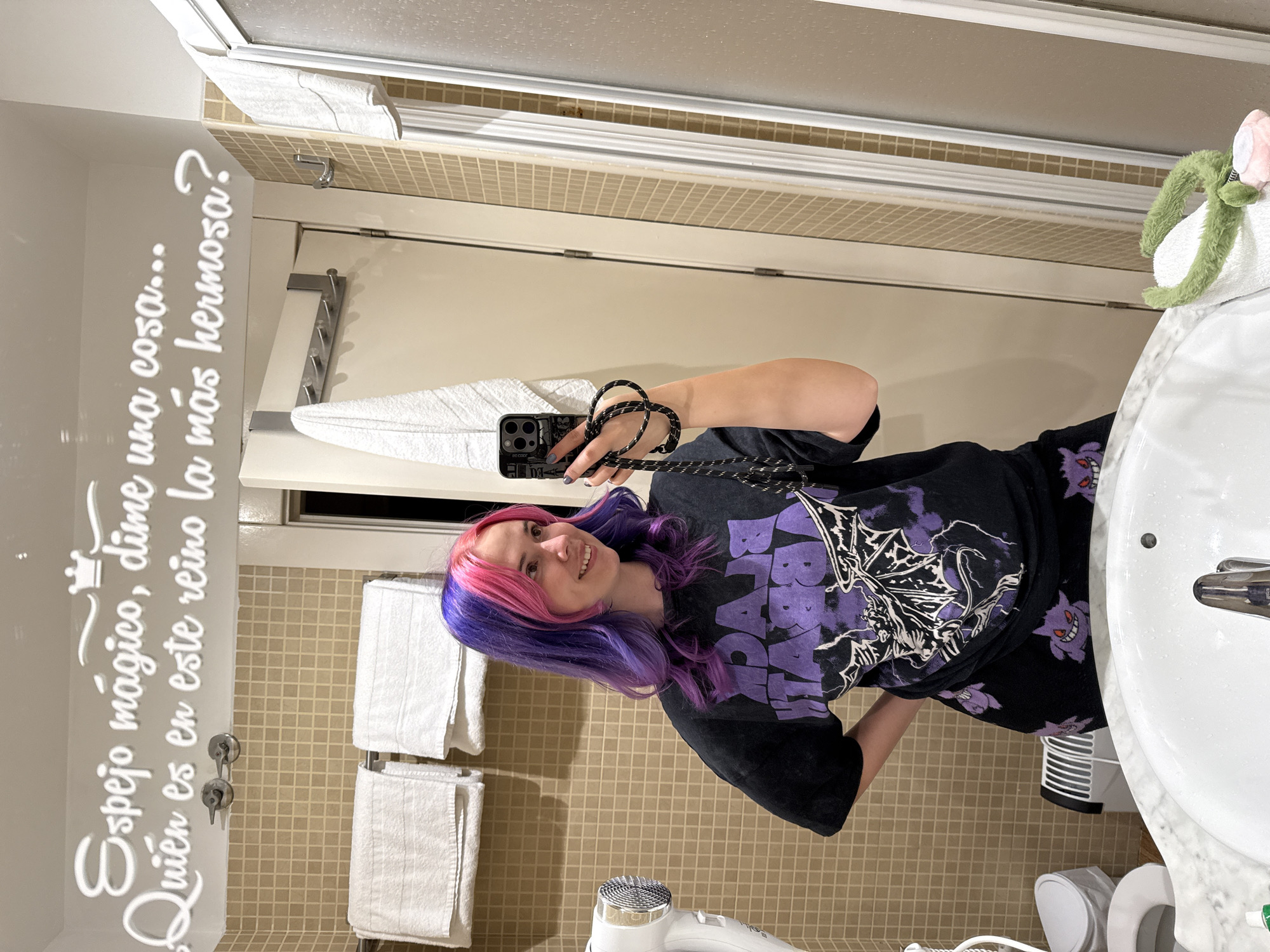A young woman with pink and purple hair is taking a selfie in a bathroom. She is holding her phone up with her right hand, which has black nail polish. She is wearing a black t-shirt with a purple Black Sabbath graphic on it, and patterned black bottoms. The bathroom has beige tiled walls and white towels hanging on the wall. The mirror has text on it in Spanish "Espejo mágico, dime una cosa... Quién es en este reino la más hermosa?". There is a sink and toilet visible. The location is a bathroom; there is no indication of the location of Madrid in the image.