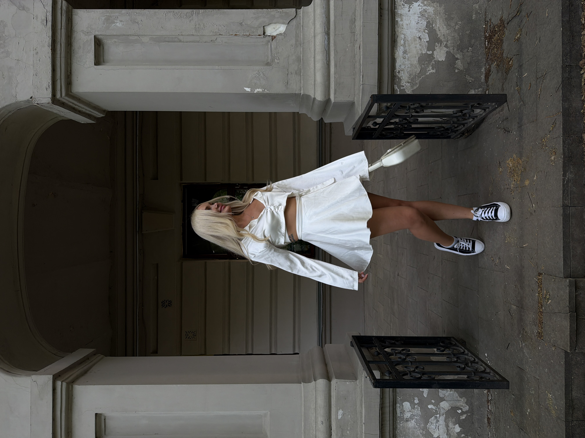 A woman with long blonde hair is posing in front of a building in Kazan, Russia. The woman is wearing a white, two-piece outfit and black and white sneakers. She has a white purse on her right shoulder. She is standing on a stone floor next to a black wrought-iron fence. The building is made of stone and has arched doorways. The overall scene appears to be an urban area.