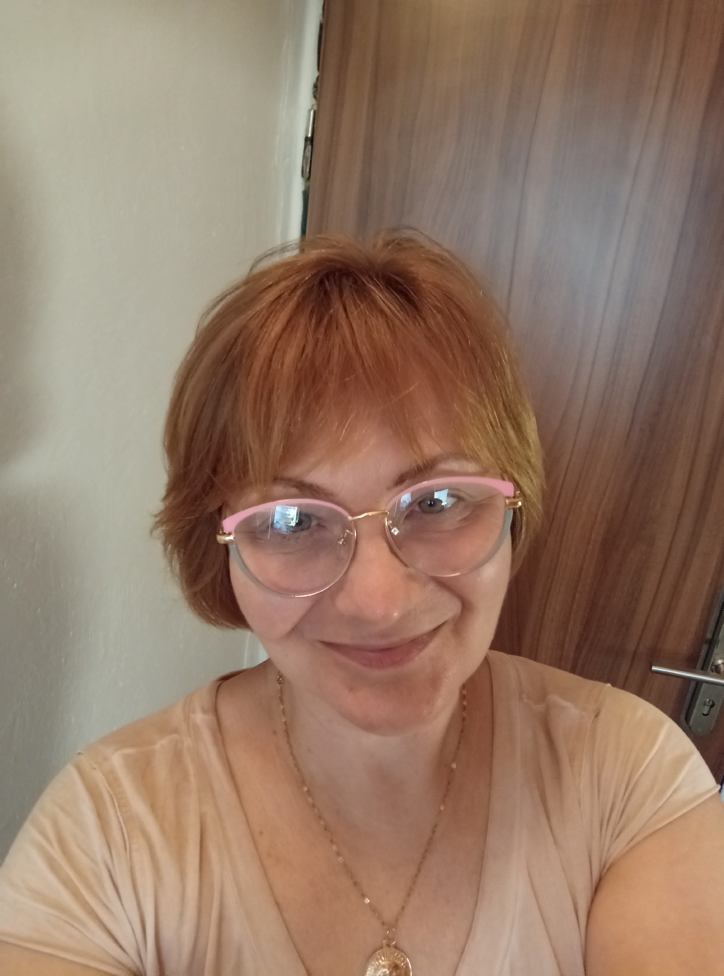 A mid-shot of a Caucasian woman with reddish-brown hair and eyeglasses is presented. She is wearing a light-colored t-shirt and a necklace with a pendant. The background consists of a plain wall on the left and a wooden door with a door handle on the right. The scene appears to be indoors, possibly in a residential setting. There are no discernible landmarks that would indicate a specific location within Brno, Czechia. The lighting is even, suggesting artificial illumination. The woman's facial expression is neutral, and her gaze is directed towards the camera.