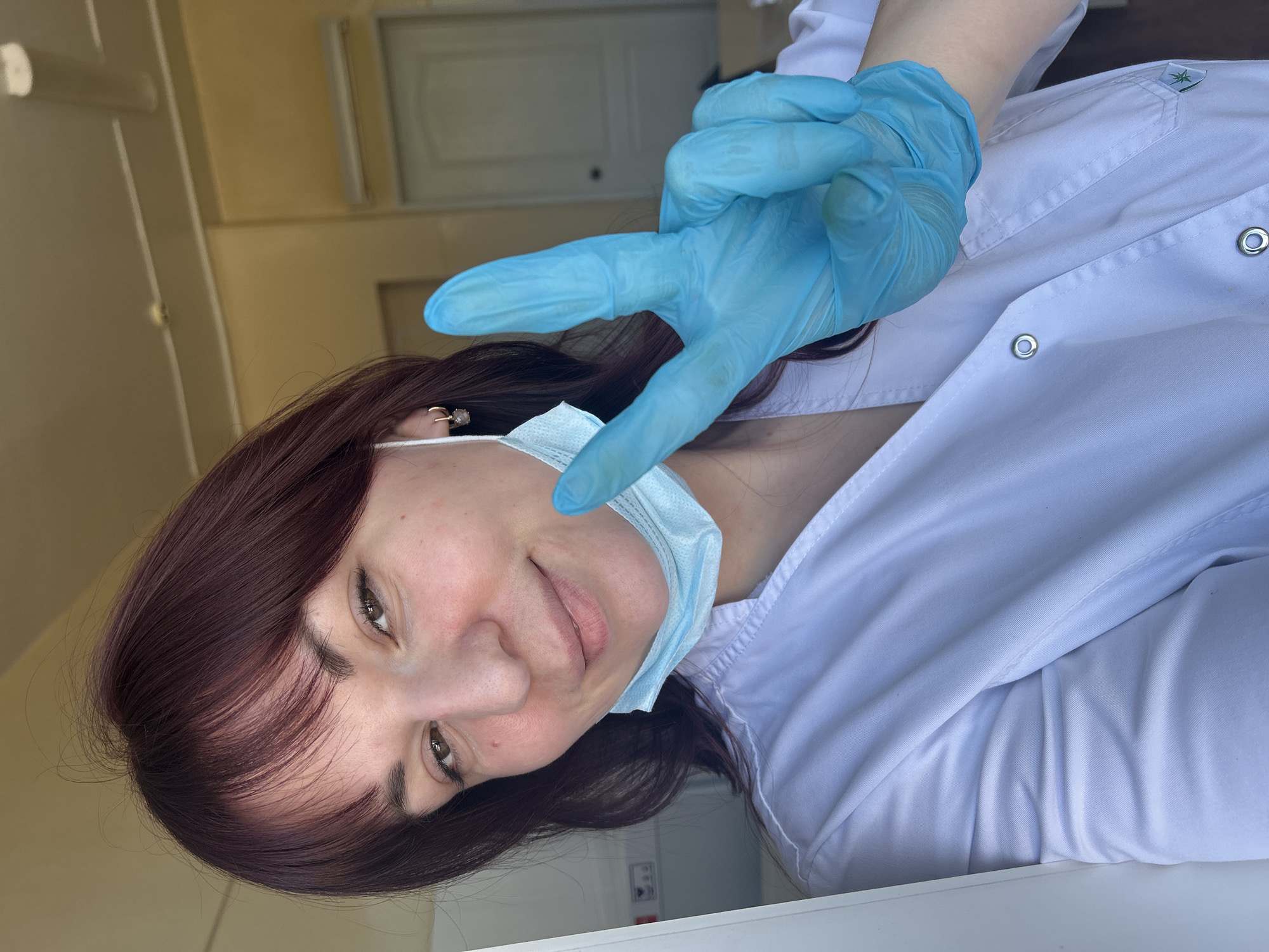 A woman in a white medical coat and blue gloves is making a peace sign with her gloved hand. She is also wearing a blue surgical mask, which is hanging off of her face. The location appears to be indoors, possibly a medical facility, as suggested by the medical coat and mask. The city is Фоки, Russia.