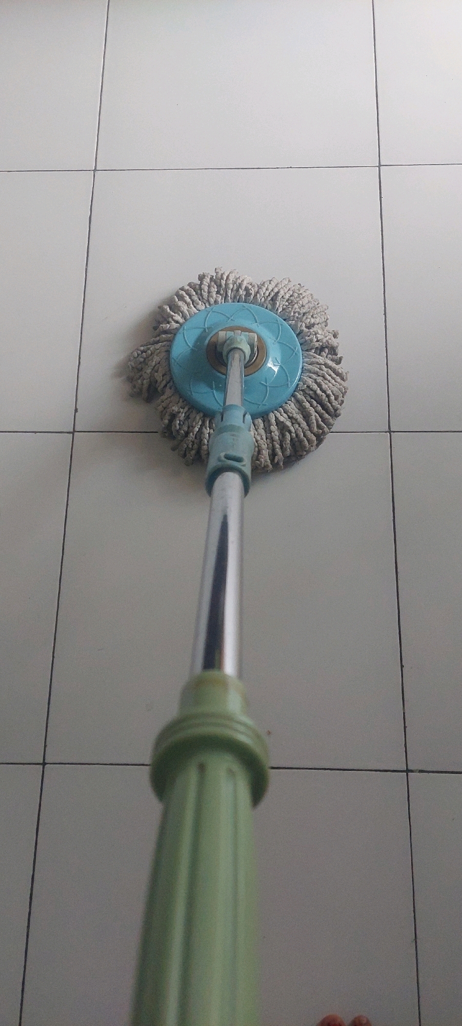 An eye-level, medium shot shows a spin mop on a tiled floor. The mop head is light brown and fluffy, with a blue plastic circular component in the center. A silver pole extends from the mop head, connected to a ribbed green handle at the bottom of the frame. The floor is covered in large, light gray square tiles with thin dark grout lines. The image is taken indoors. The exact location within Lumajang, Indonesia, cannot be determined from the visual information provided.