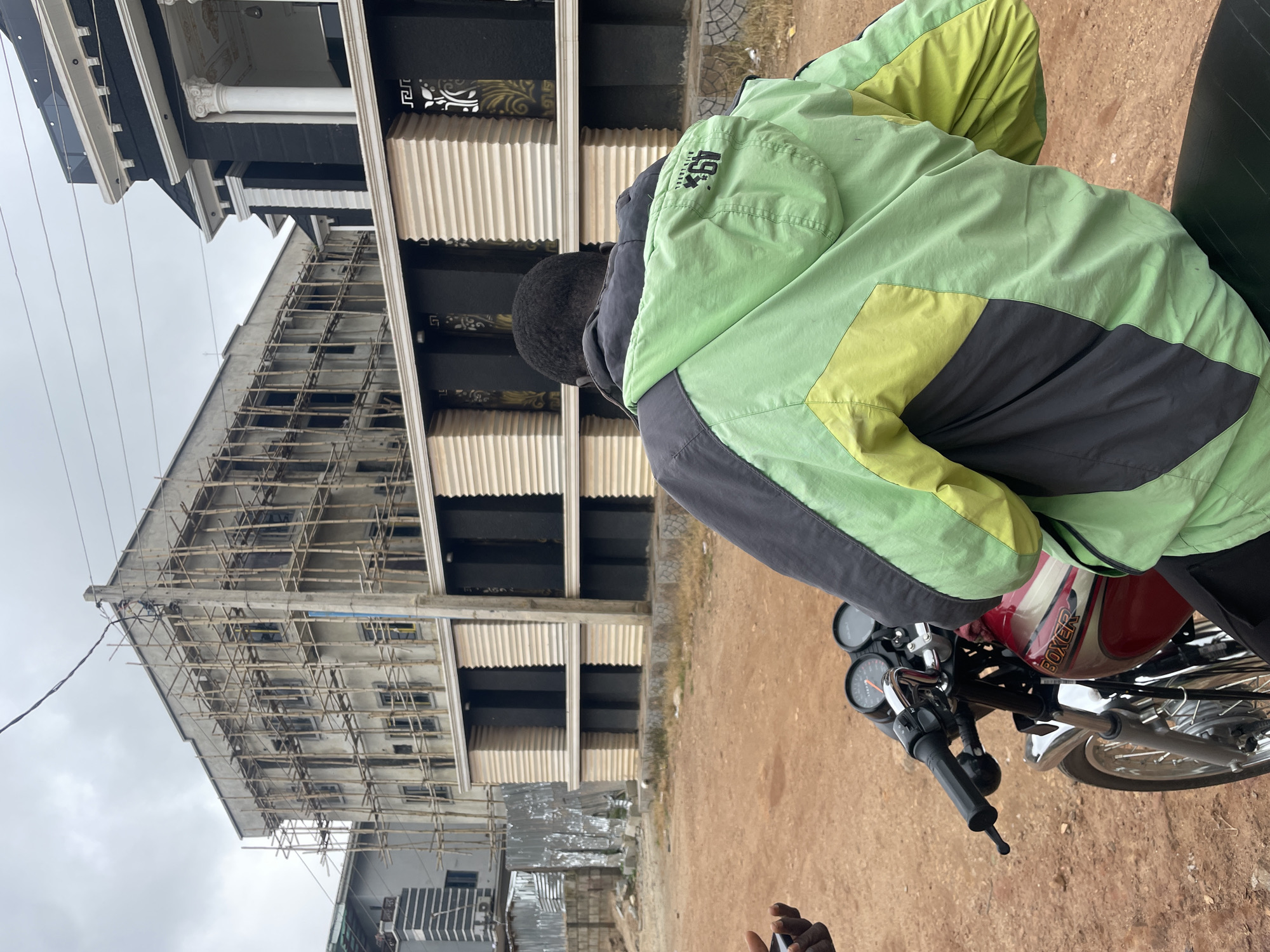 An overhead view captures a man from behind, seated on a red motorcycle. He wears a vibrant green, yellow, and dark grey jacket. The motorcycle's dashboard features two circular gauges. The man's left hand, holding a mobile phone, is visible in the lower left corner of the frame.
In the background, a multi-story building is under construction, shrouded in bamboo scaffolding. Adjacent to it, a partially completed modern building displays a black facade with white ornate columns and decorative gold patterns. A low wall with horizontal ribbed panels in cream and dark colors separates the street from the buildings. The ground is a dusty, reddish-brown earth, and the sky above is overcast. The scene appears to be in Abuja, Nigeria, given the architectural style and construction methods.