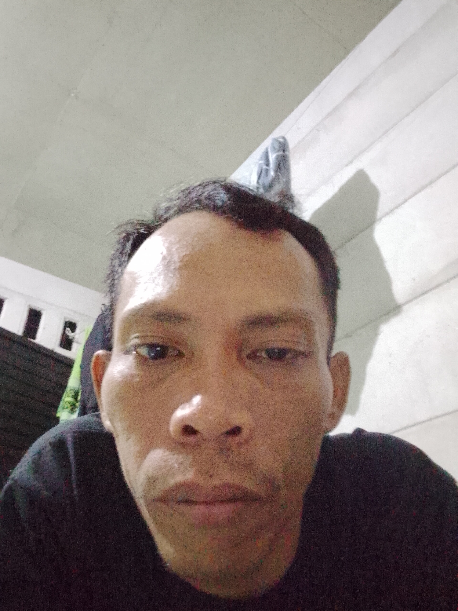 A headshot of a man with dark hair, brown skin, and a black shirt, taken from a low angle. The background consists of a white painted wall with vertical grooves on the right side of the image, and a white ceiling above. In the background on the left, there is a dark-colored cabinet and a partly visible green shirt. The location is Bukit Ketok, Indonesia.